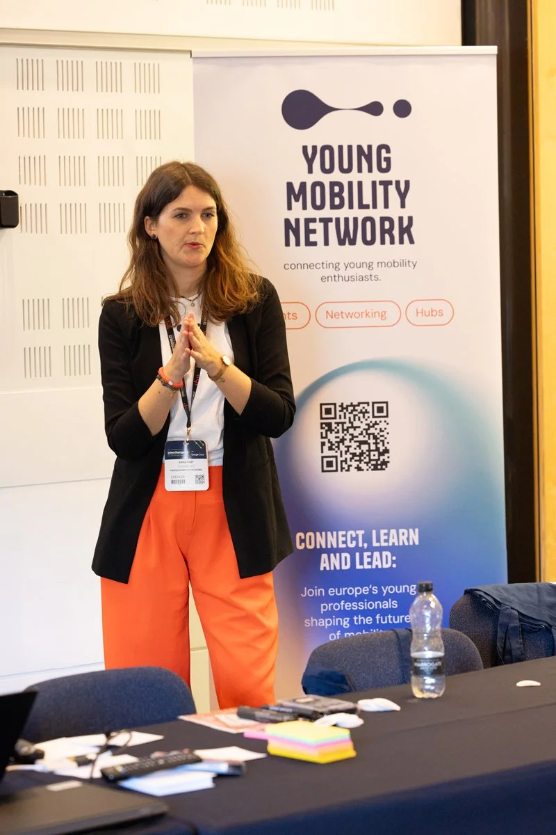 A woman standing in front of a banner for the Young Mobility Network, which features a logo with a stylized shuttle and text promoting connecting young mobility professionals. The woman appears to be speaking or presenting, with her hands clasped tog