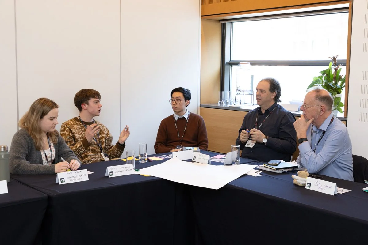 Five people seated around a conference table engaged in a discussion, with notebooks, glasses of water, and name tags in front of them.