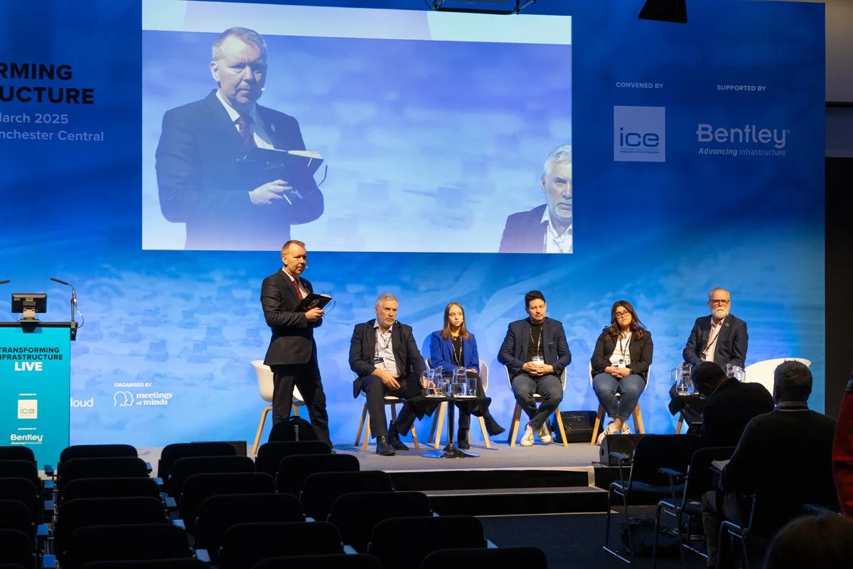 A panel of six people on stage during a conference, with one standing and speaking into a microphone, while the others are seated. A large screen behind them displays a close-up of the standing speaker. The background features blue branding with logos and text related to the event.