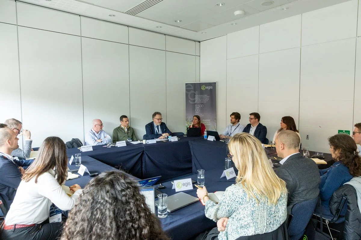 A group of people sitting around a U-shaped conference table in a modern conference room, engaged in a meeting or panel discussion.
