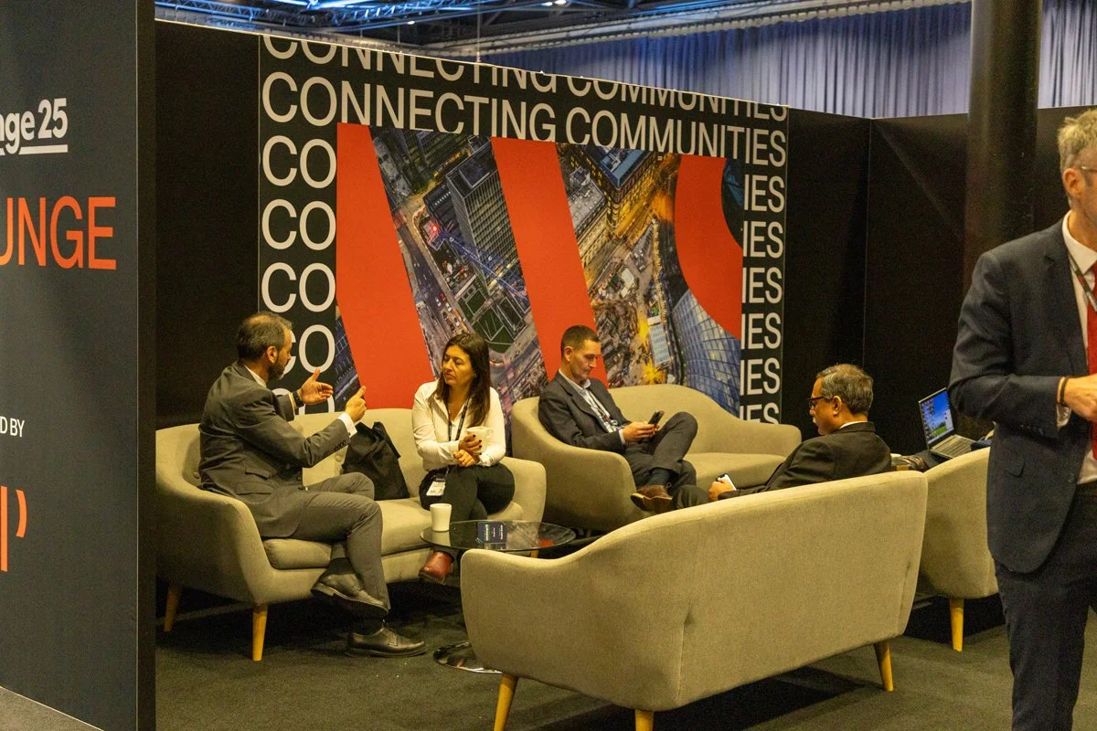 Business professionals sitting and talking on gray sofas at a conference or event, with a black backdrop featuring the words 'Connecting Communities' and a cityscape graphic.