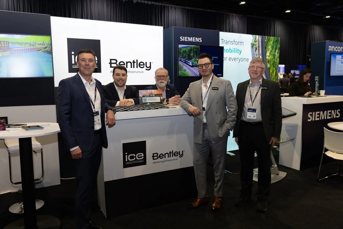 Group of five men in business attire standing and sitting at a Bentley-themed exhibit booth with Siemens branding at a conference or trade show.
