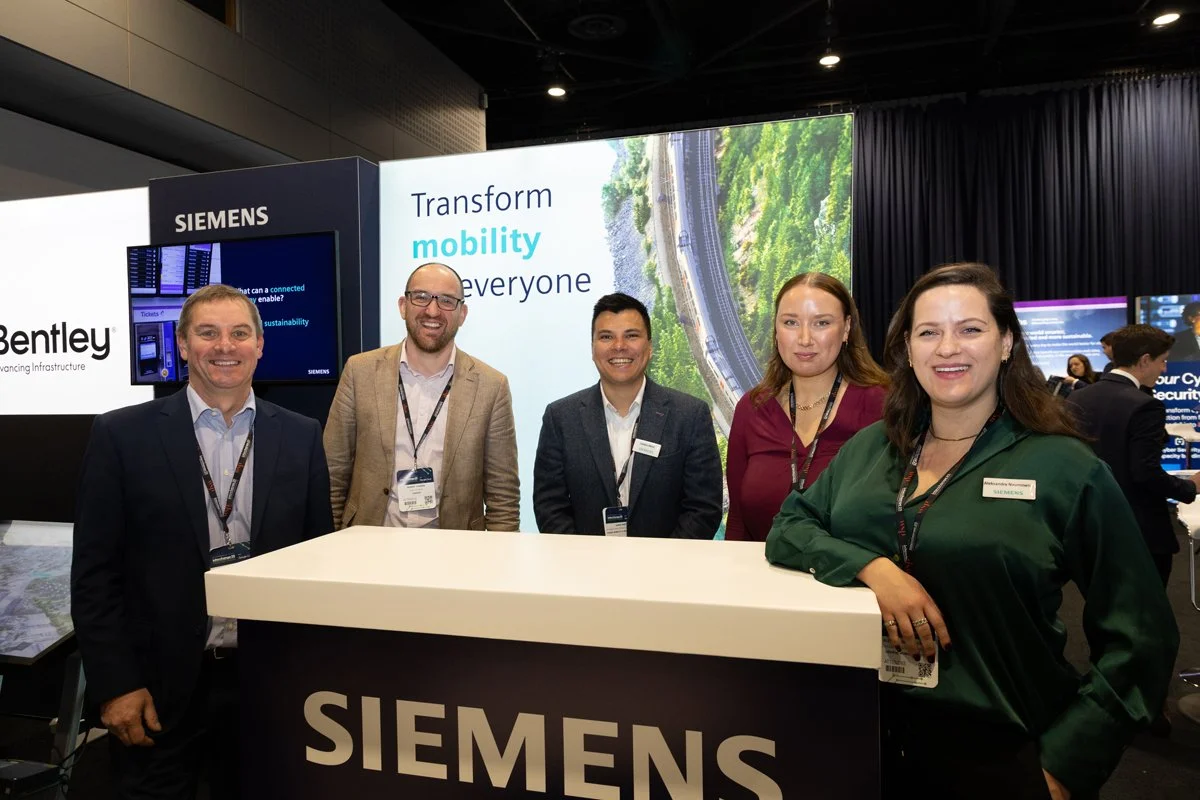 Group of five smiling professionals at a Siemens booth during a corporate event, with a large digital screen in the background displaying the words "Transform mobility for everyone."