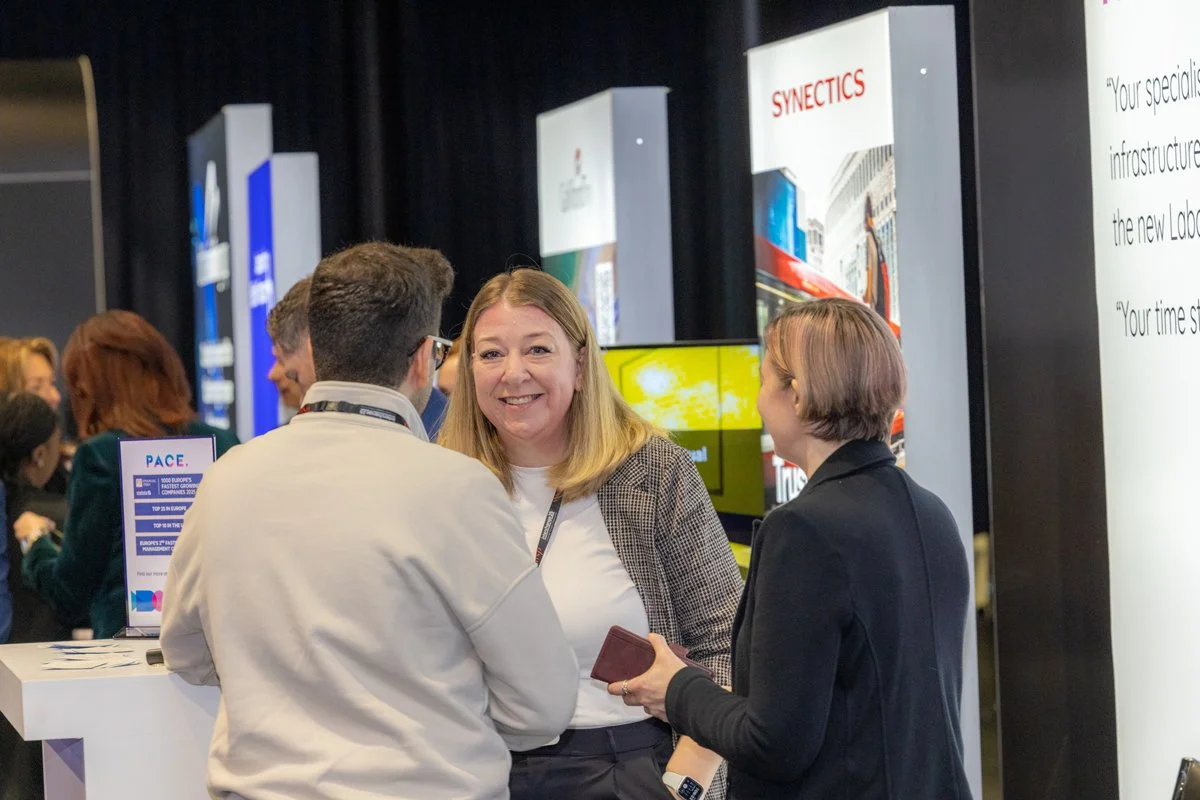 Three people engaged in conversation at a professional event or conference, with booth displays and banners in the background.