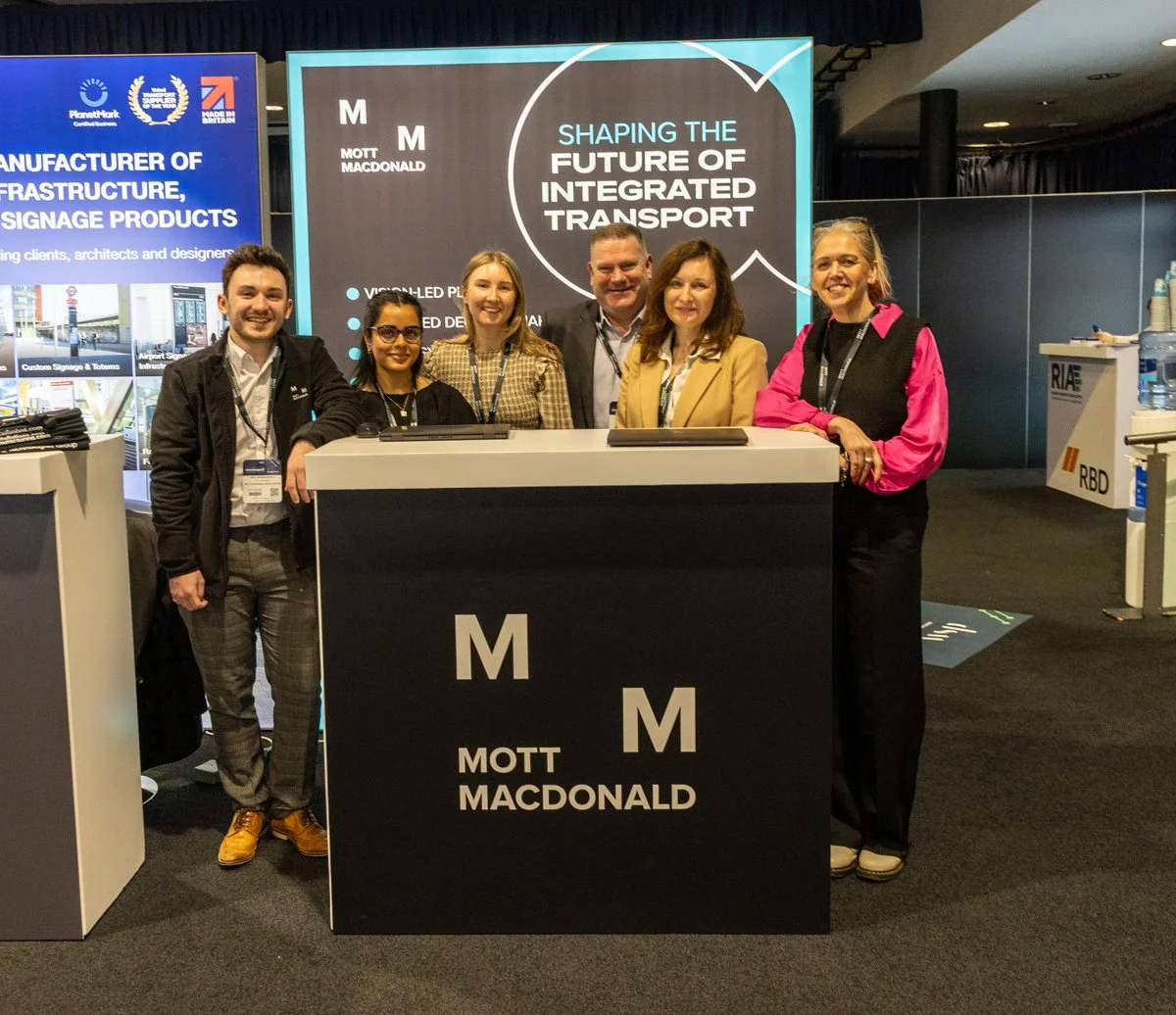 A group of six people standing behind a booth at a conference or trade show, with a large display behind them that reads "Shaping the Future of Integrated Transport."