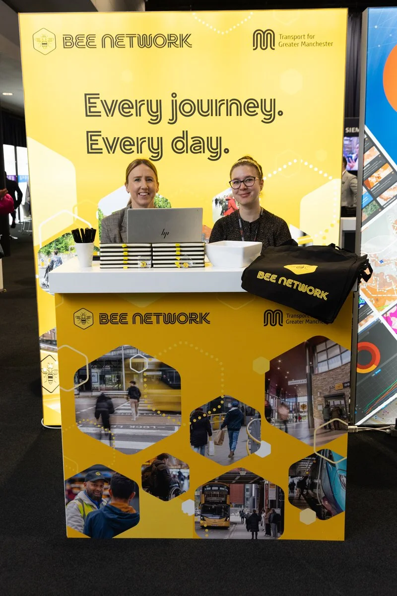 A promotion booth for BEEl NETWORK and Transport for Greater Manchester features a bright yellow background with the slogan "Every journey. Every day." Two women are sitting at the booth, smiling. The booth displays images of pedestrians and public t