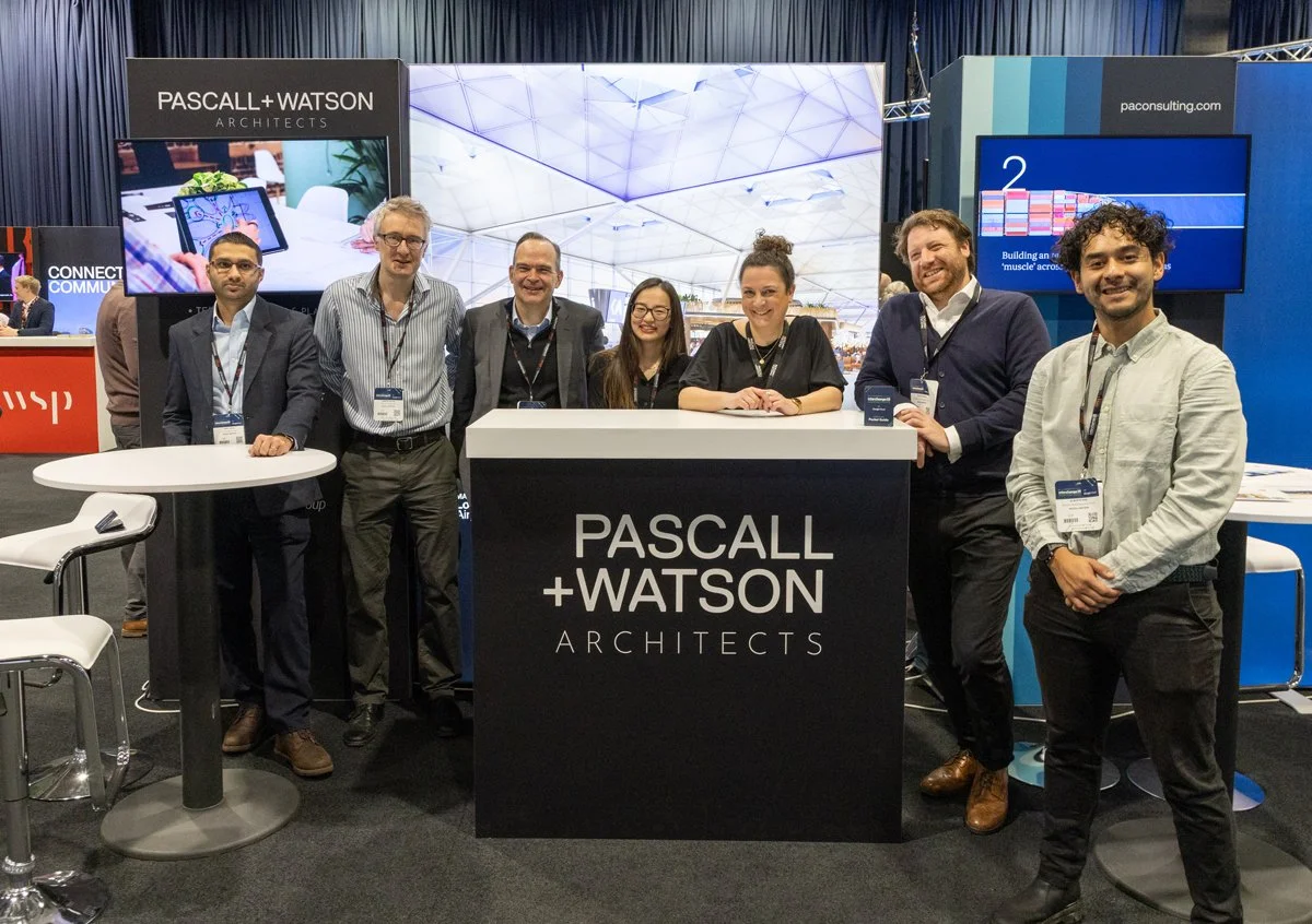 Group of seven people standing behind a booth with a sign that reads "PASCALL + WATSON ARCHITECTS" at a trade show or conference.