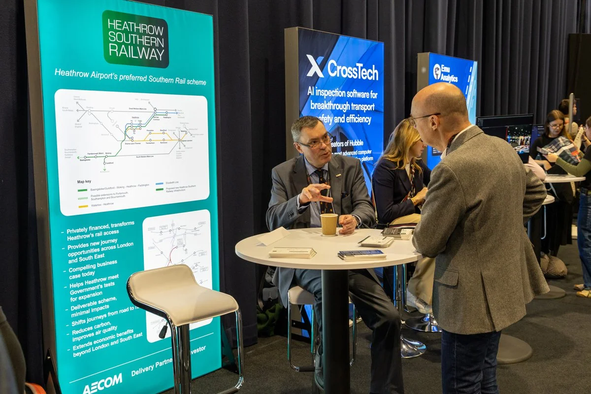 Business person in a gray blazer and glasses talking to another man with glasses and a gray blazer at a conference booth with display boards about Heathrow Southern Railway and CrossTech.