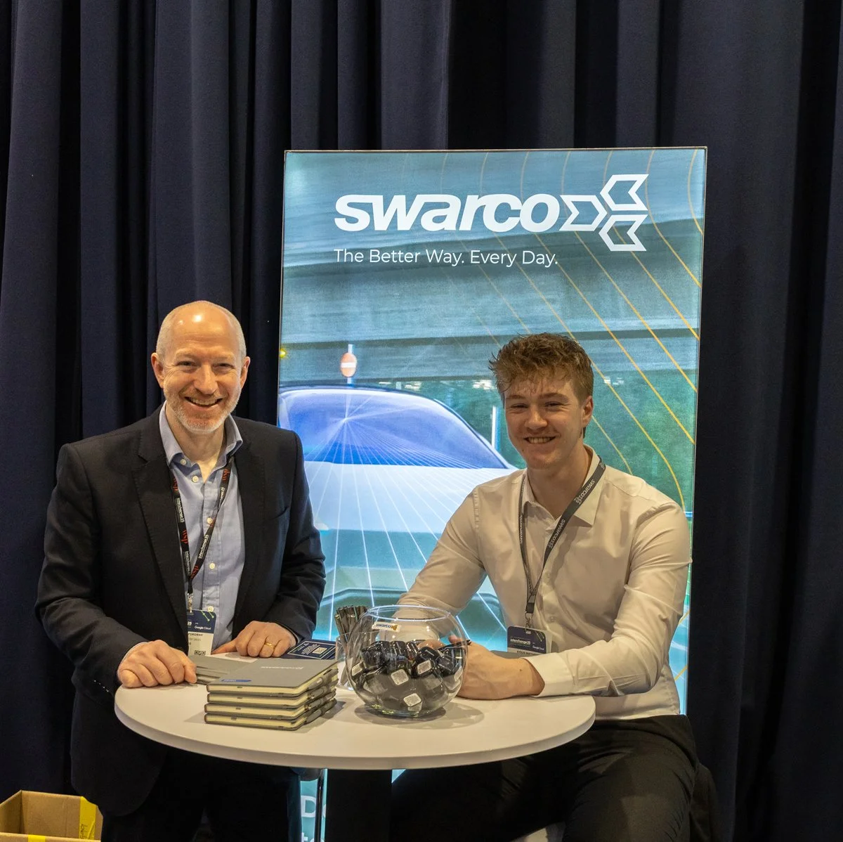 Two men smiling at a booth with a Swarco banner in the background, one opened a book on a small round table with a bowl of black items, in a professional event setting.