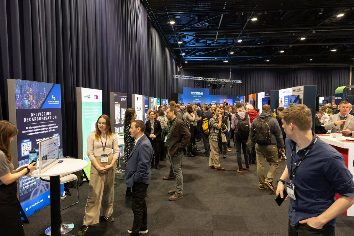 Crowd of people at a conference or exhibition hall with various display banners and booths.