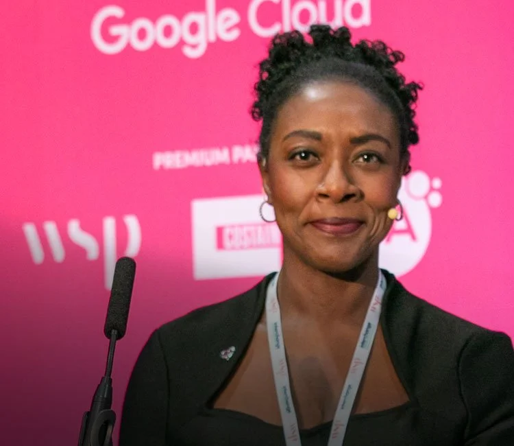 A woman with short curly hair, wearing a black blazer and a white lanyard, standing in front of a pink backdrop with the words 'Google Cloud' and other logos, smiling slightly.