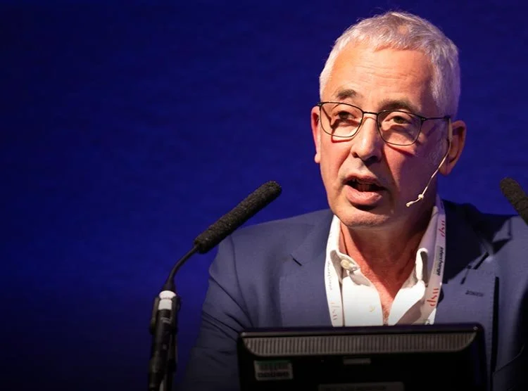 A middle-aged man with gray hair and glasses speaking at a conference, wearing a blue suit and white shirt, with a dark blue background and two microphones in front of him.