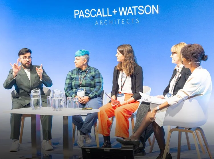 Panel discussion at Paschall + Watson Architects conference with five people seated on stage, engaging in conversation, with a blue backdrop displaying the company name.
