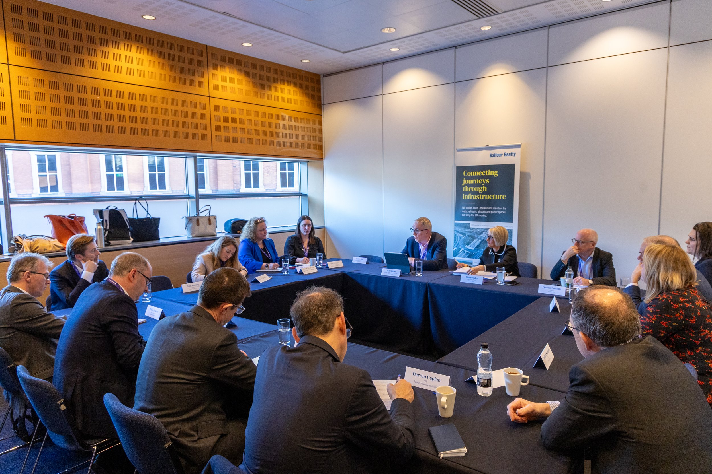 A professional conference or meeting taking place indoors, with attendees seated around a large rectangular table. Several people are engaged in discussion, with name tags, notebooks, and cups on the table. A standing banner with the title 'Connecting journeys through infrastructure' is visible in the background.