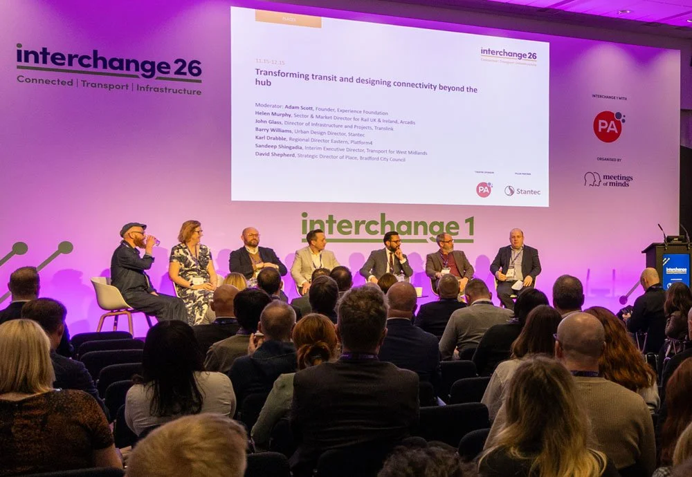 Panel discussion at the exchange26 conference with seven speakers seated on stage, a large screen behind them displaying a presentation titled 'Transforming transit and designing connectivity beyond the hub,' and a seated audience watching.