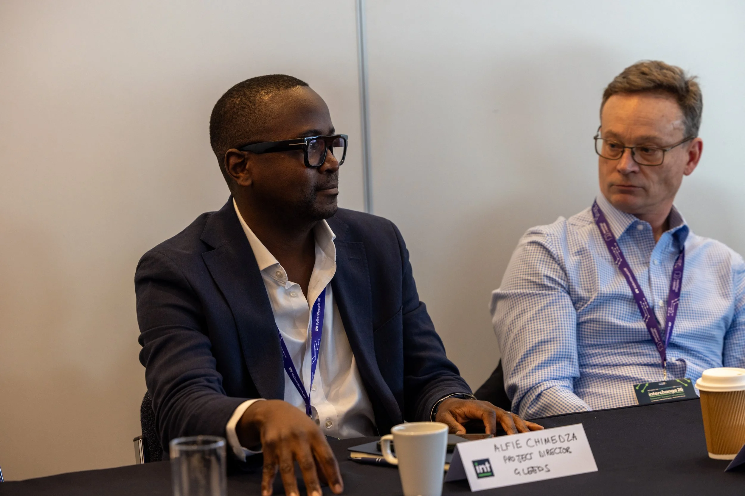 Two men sitting at a conference table, engaged in discussion. The man on the left is wearing glasses and a dark suit, while the man on the right is wearing glasses and a checked shirt. There are nameplates, coffee cups, and conference materials on the table.