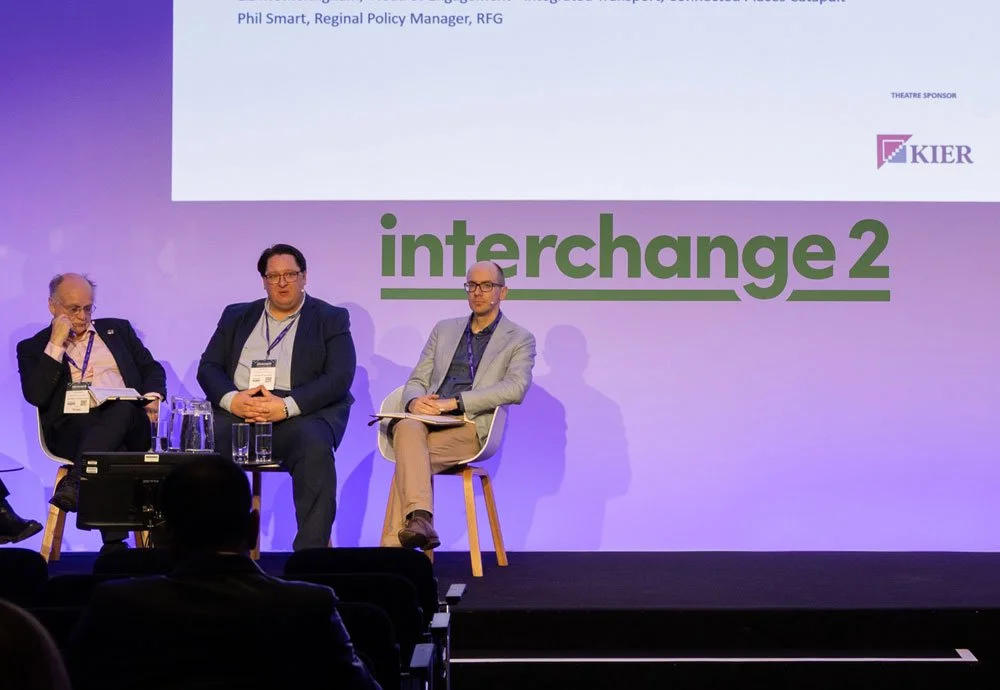 Three men sitting on a stage at a panel discussion, with a large screen behind them displaying the words "interchange 2" and a logo for KIER. Audience members are visible in the foreground.