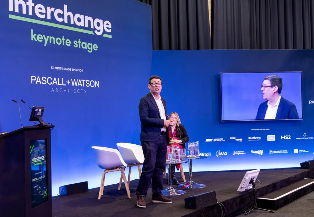 A man in a dark suit speaking on stage at the Interchange conference, with a woman seated behind him, and the conference branding on a blue backdrop. There is a large screen displaying the speaker and a podium to the side.