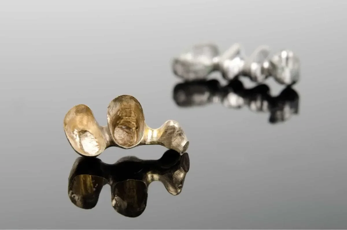 Two metallic bean-shaped objects with one in gold tone in the foreground and one in silver tone in the background, both reflected on a glossy black surface.
