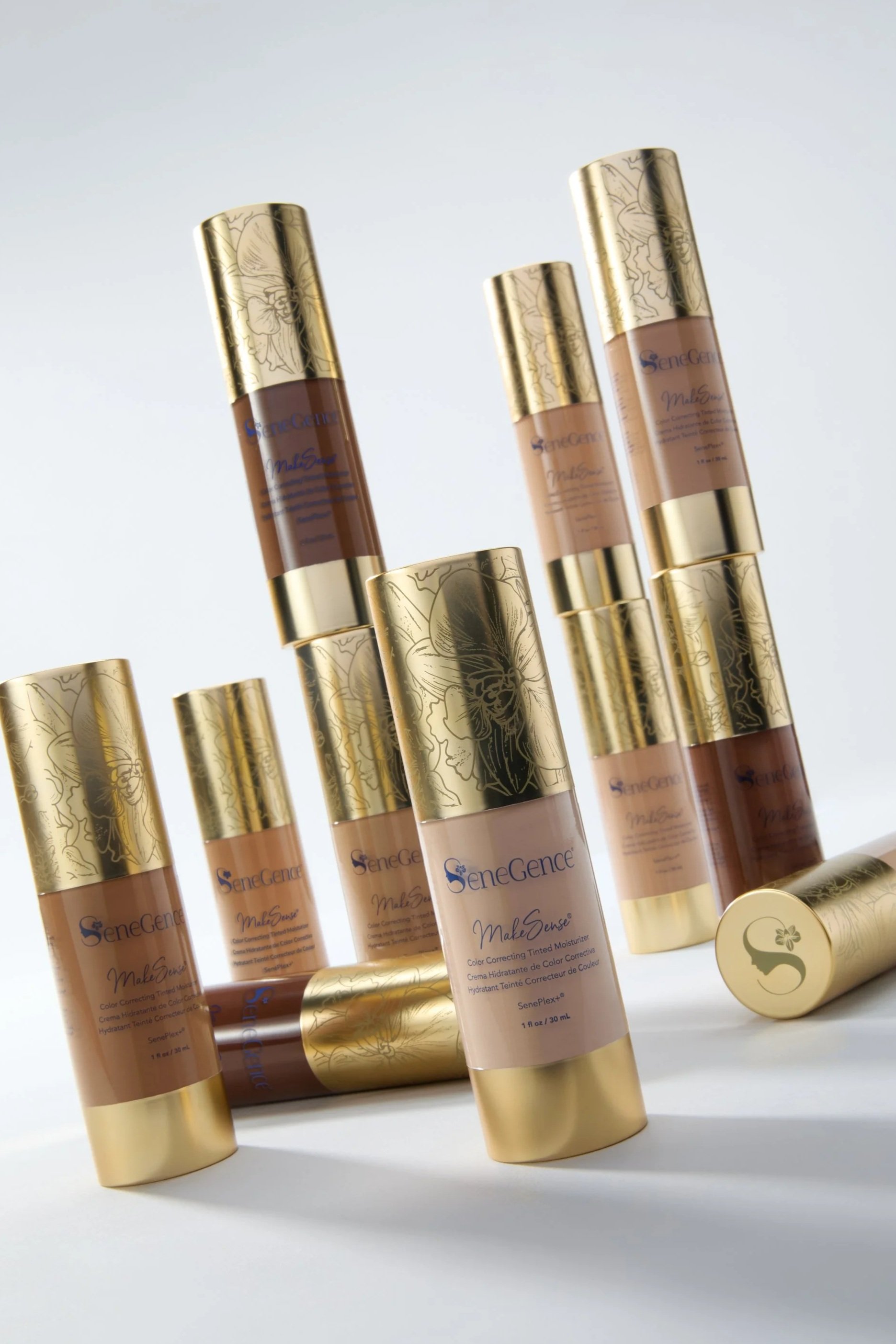 Multiple tubes of SeneGence MakeSense color correcting tinted moisturizer with gold caps and labels featuring floral designs, arranged on a white surface.