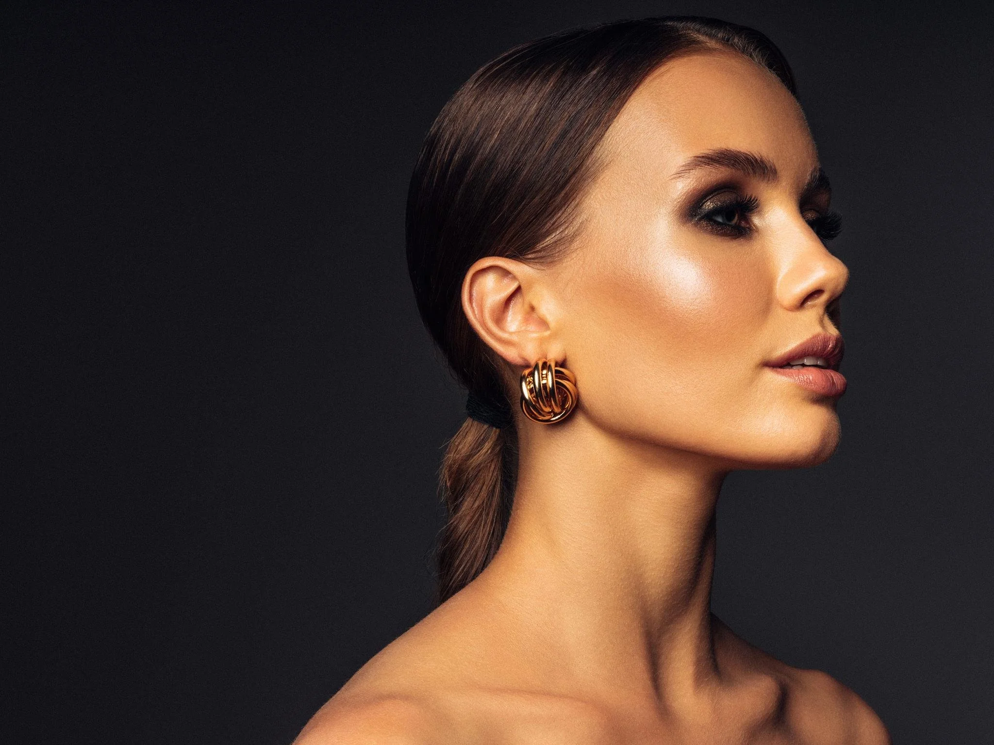 Close-up of a woman with dark hair slicked back, wearing large gold earrings, gold eye makeup, and soft pink lipstick, against a black background.