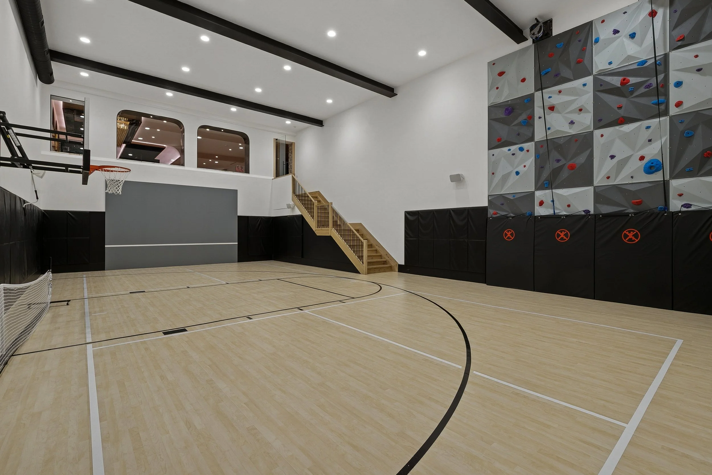 Indoor gym with a wooden basketball court, climbing wall, and a staircase, illuminated by ceiling lights.