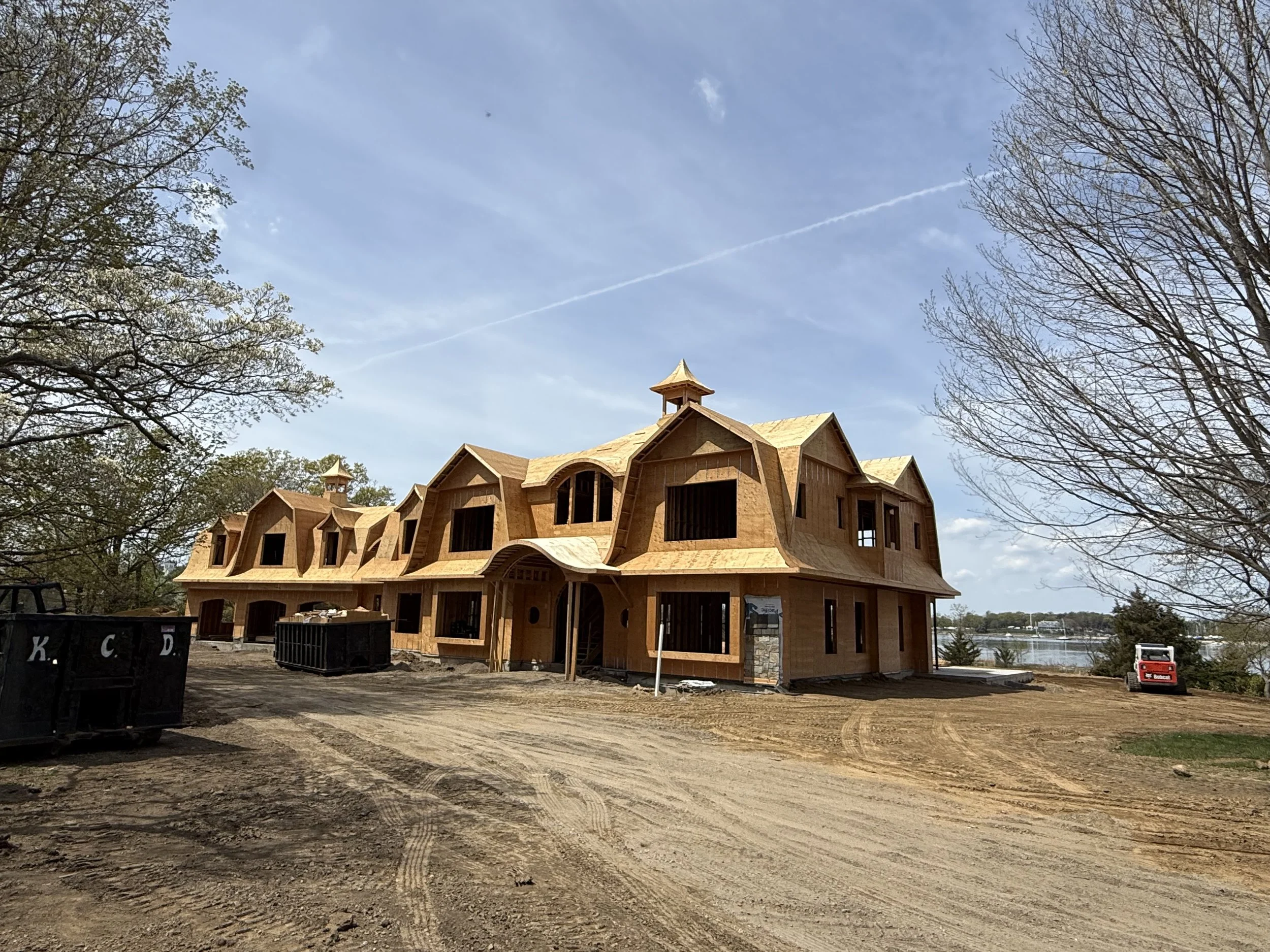 Under construction house with wooden framing and steep roof, surrounded by trees and construction equipment, with a clear blue sky.