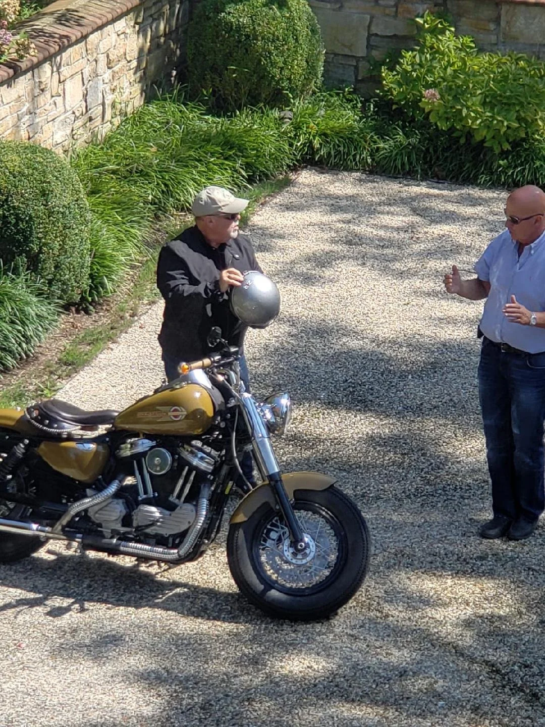 Two men talking outdoors with a vintage gold motorcycle parked nearby. One man wears a black jacket, sunglasses, and a beige cap, holding a helmet. The other man wears a white shirt and jeans, gesturing with his hands. They stand on a gravel path surrounded by green bushes and a stone wall.