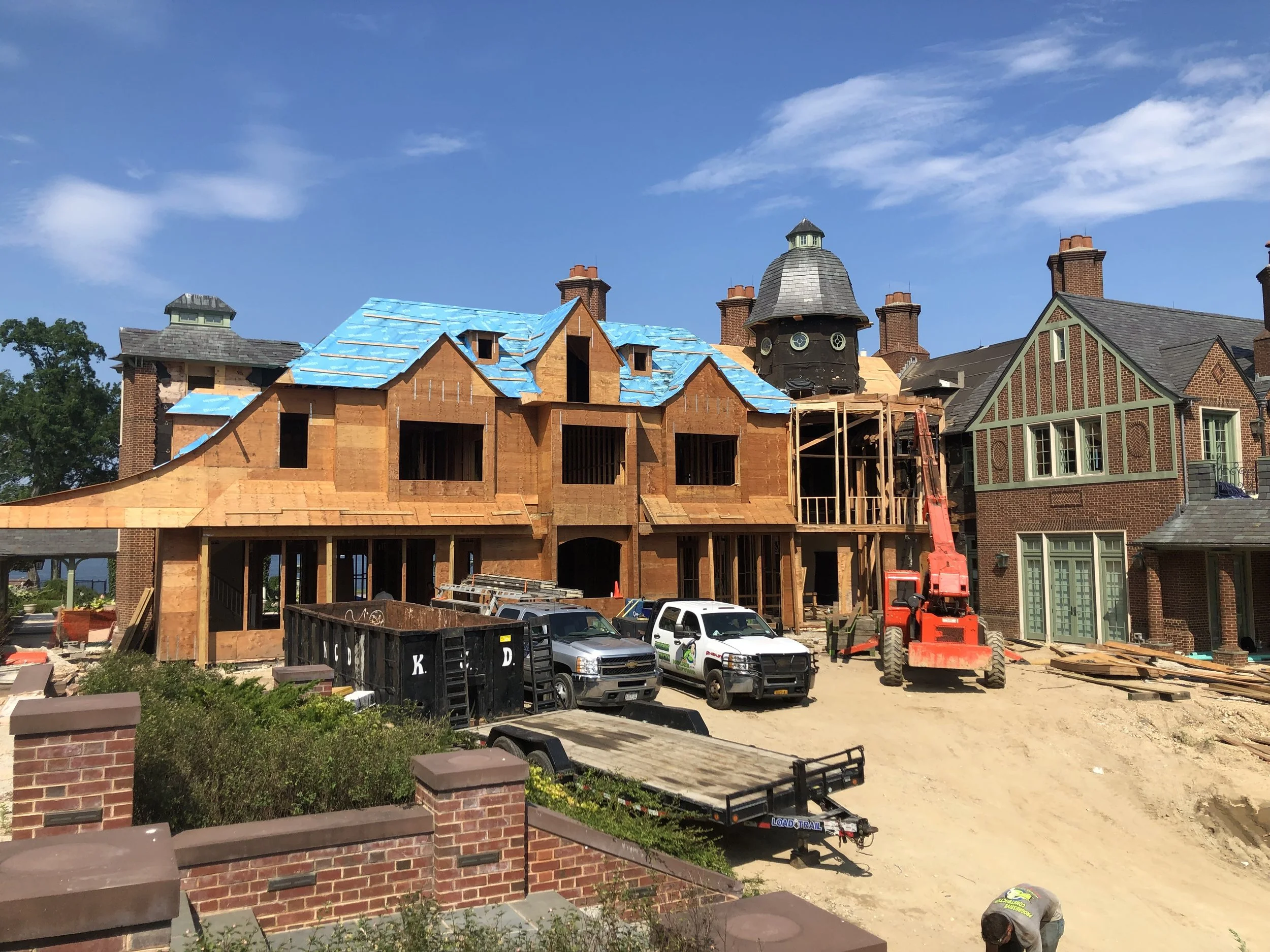 Residential house under construction with blue roofing, surrounded by construction vehicles and materials, and a clear blue sky.