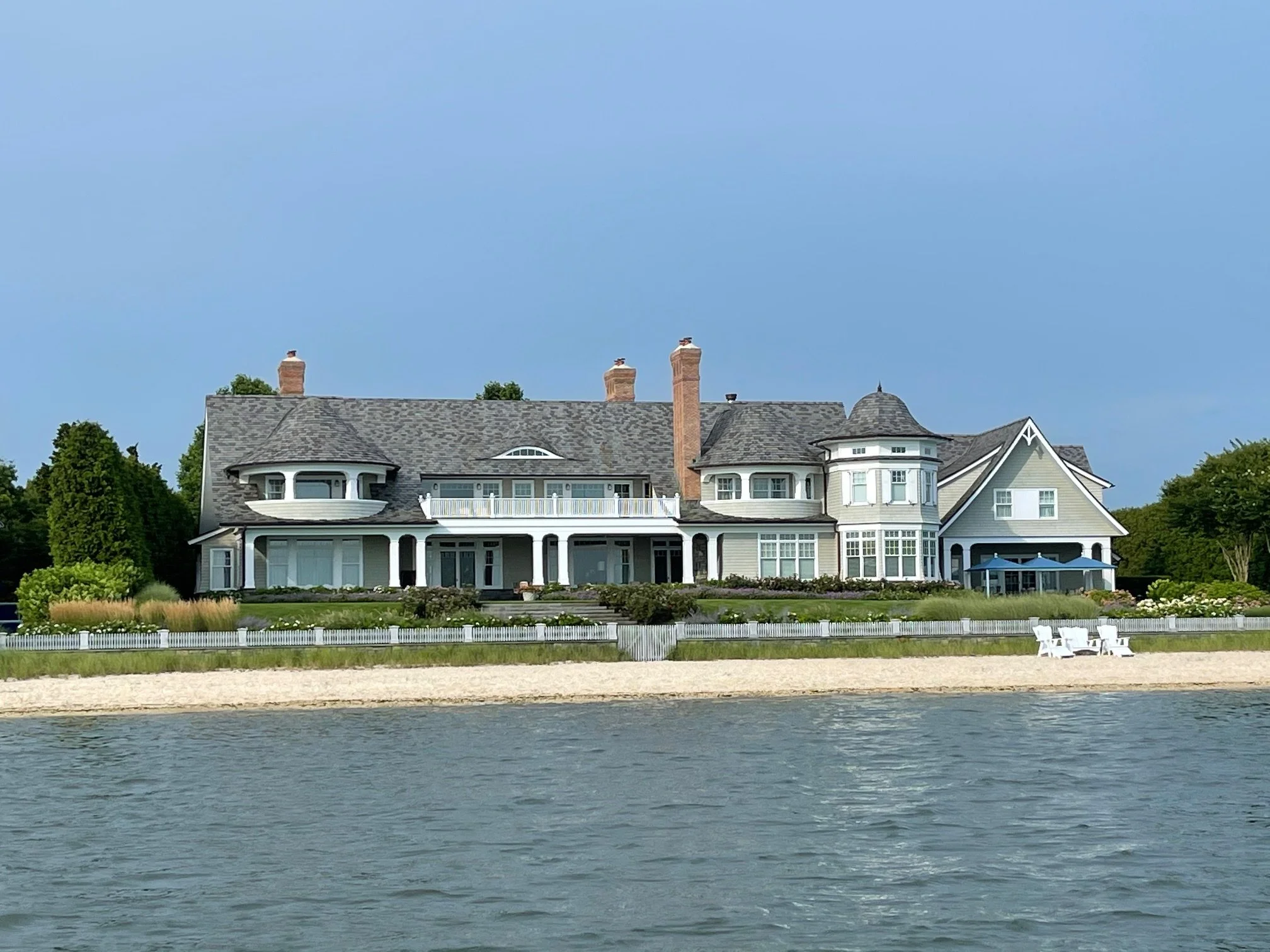 Large white house with multiple chimneys, turret, and wrap-around porch, situated by a body of water with a sandy shoreline and a white picket fence in the foreground. Clear sky above.