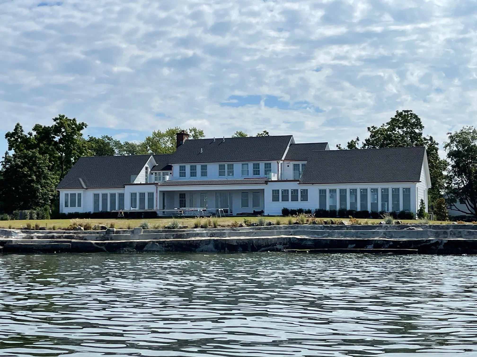 A large white house with multiple stories and a sloped black roof, situated along a body of water with a rocky shoreline. Trees are visible behind and around the house, and the sky above is partly cloudy.