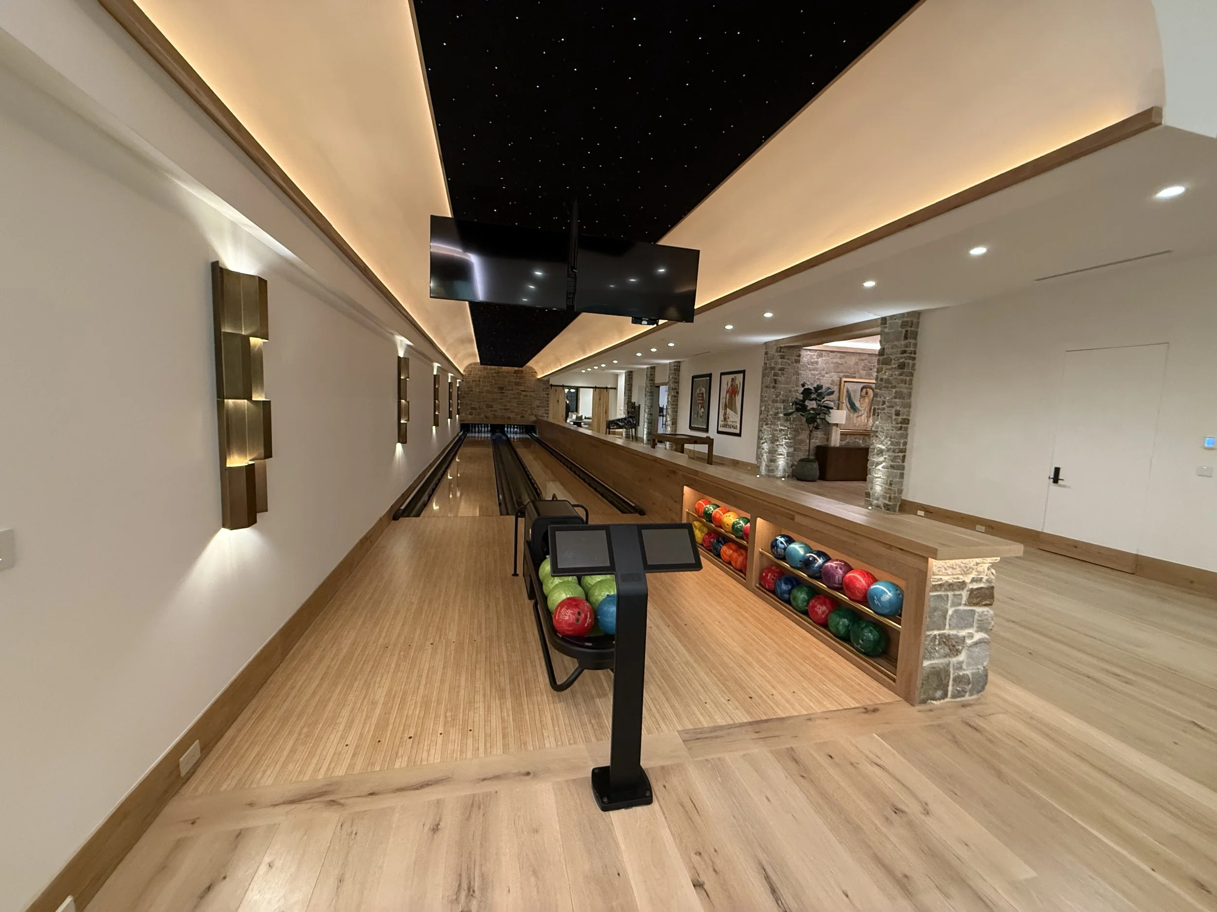 Indoor bowling alley with a lane, bowling balls, a bowling ball return system, and wooden flooring, illuminated by ceiling lights and wall sconces, with brick and painted walls