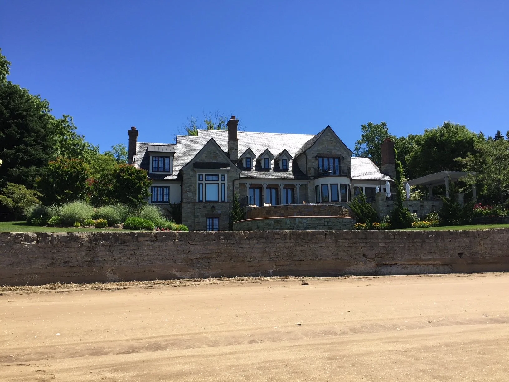 A large, elegant house with stone and white exterior, multiple chimneys, and surrounding greenery, under a clear blue sky.