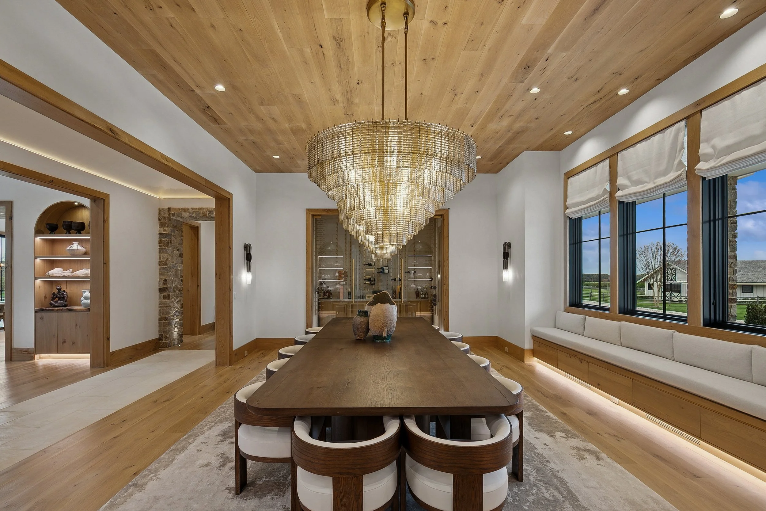 Interior of a dining room with a long wooden table, modern chairs, a large chandelier, windows with white blinds, and a built-in bench with cushions.