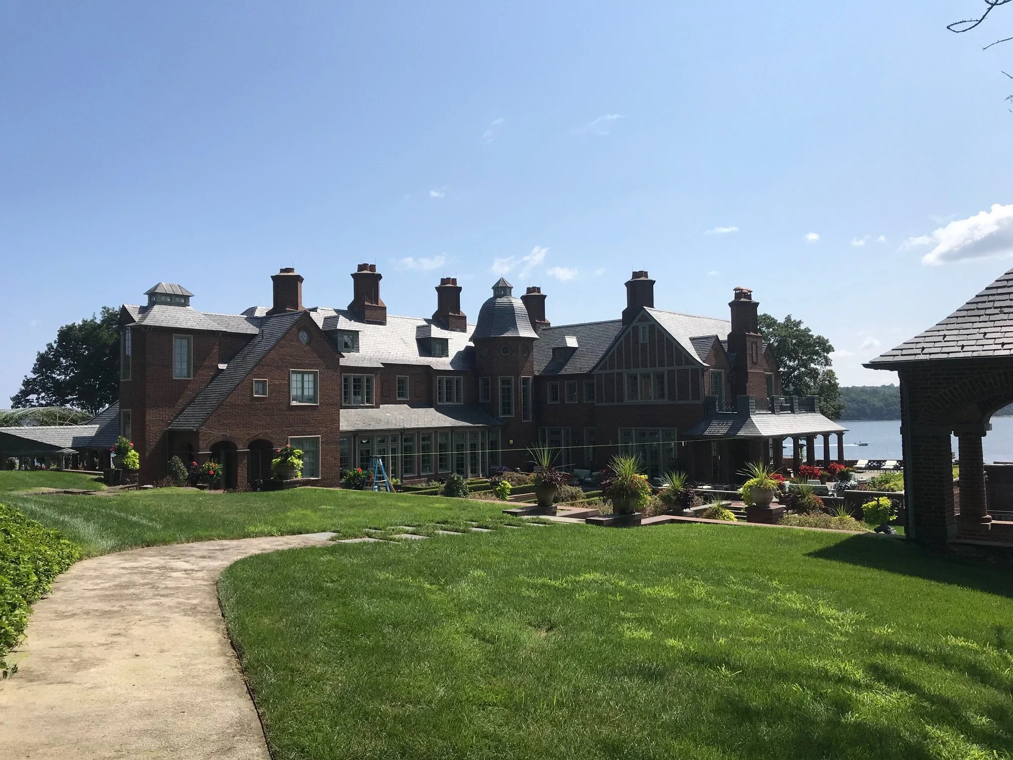 A large brick mansion with multiple chimneys, dormer windows, and a steeply pitched roof, situated by a body of water with a boat visible in the distance. The property features a well-manicured lawn with a winding stone pathway and potted plants.