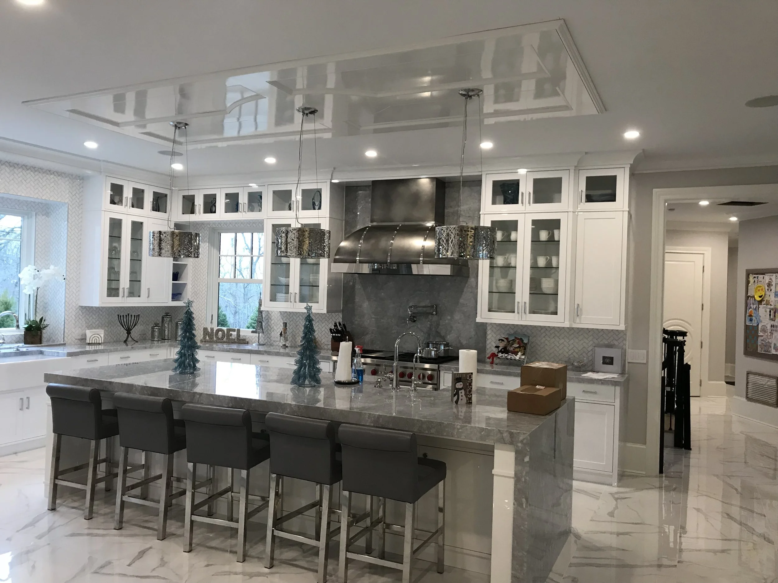 Modern kitchen with white cabinets, marble countertops, stainless steel range hood, and pendant lights with metallic patterns, decorated for Christmas with small Christmas trees and holiday decor on the island.