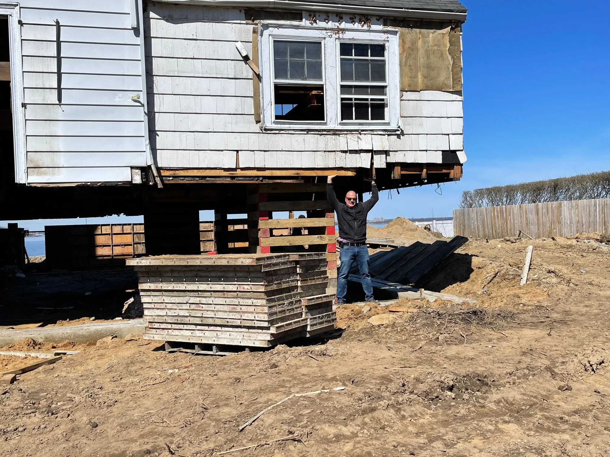 A man in sunglasses and a black jacket lifts a section of a house that is under construction or renovation, with parts of the house elevated on temporary supports on a construction site with dirt ground, an under-construction foundation, and a clear blue sky in the background.