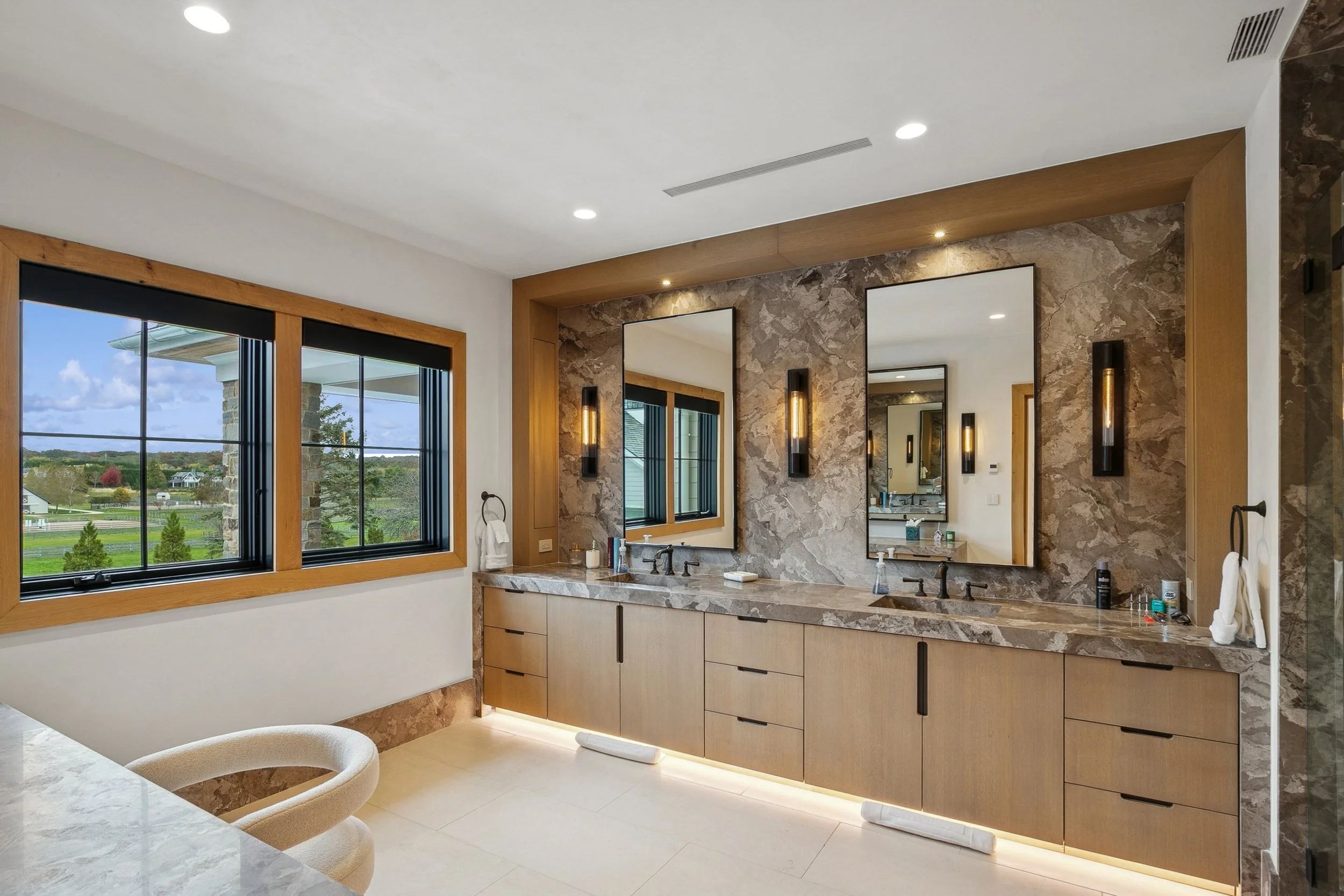 Modern bathroom with double vanity, marble countertops, large mirror, natural light from window, and stylish wall sconces, overlooking a scenic outdoor view.