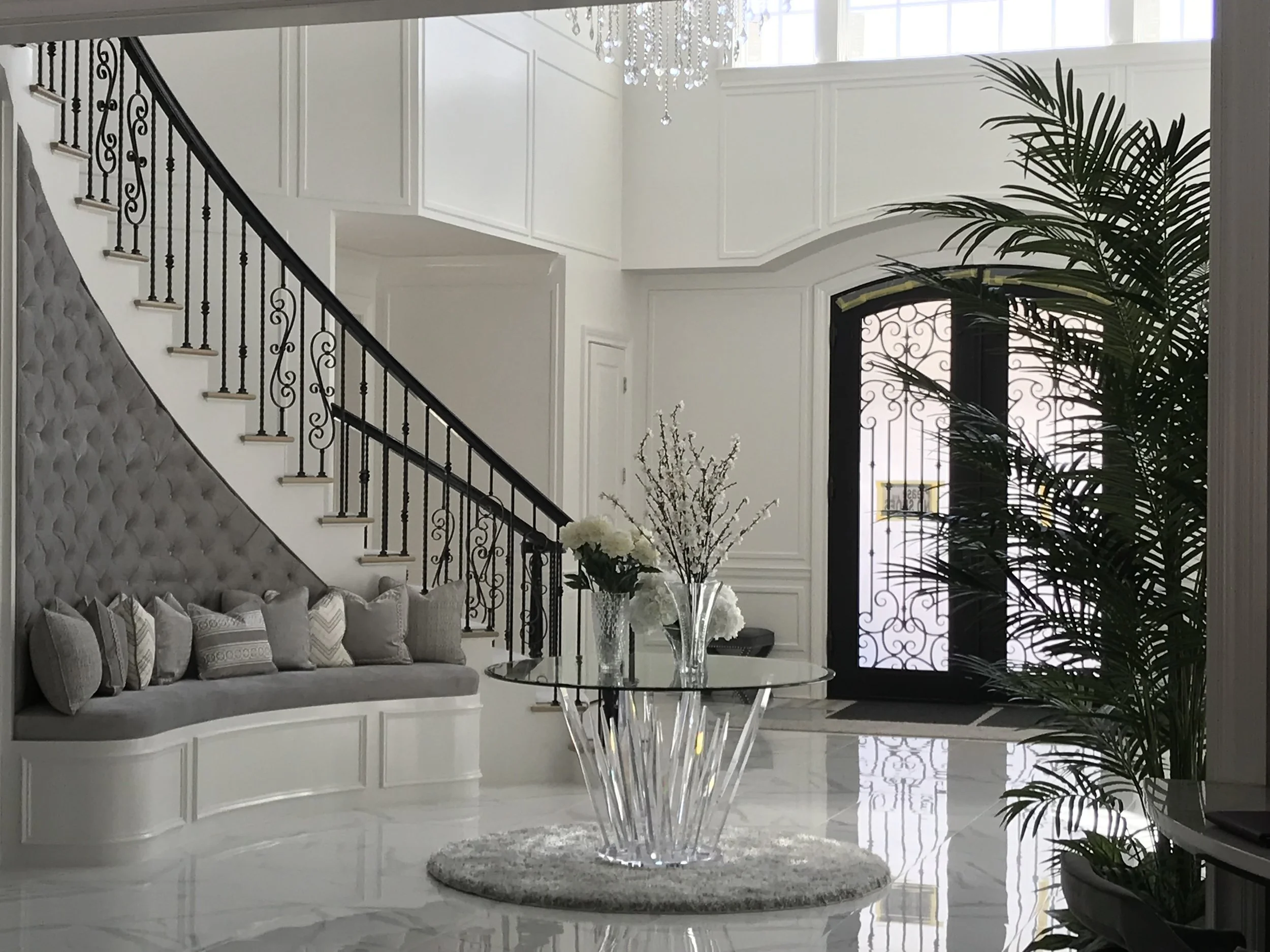 Luxurious modern living room with white walls, large window letting in natural light, a chandelier hanging from the ceiling, a black iron staircase, a grey tufted sofa with decorative pillows, a transparent glass coffee table with a flower vase, tall green plants, and a decorative front door.
