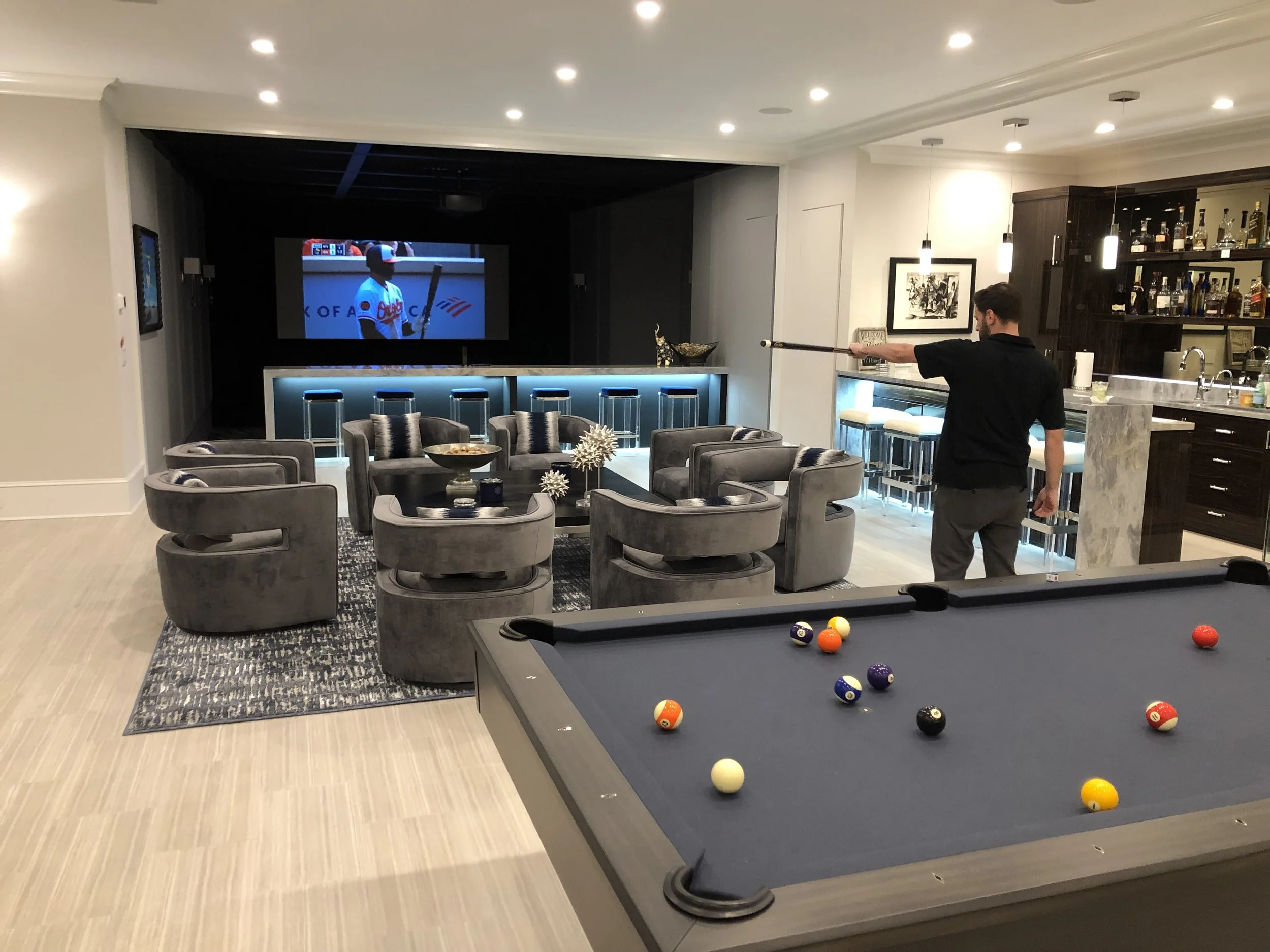 A man is playing pool on a black billiards table with colorful balls in a modern living room. There is a large TV mounted on the wall and a bar area with bottles in the background. Cozy chairs and a decorative centerpiece are arranged around the room.