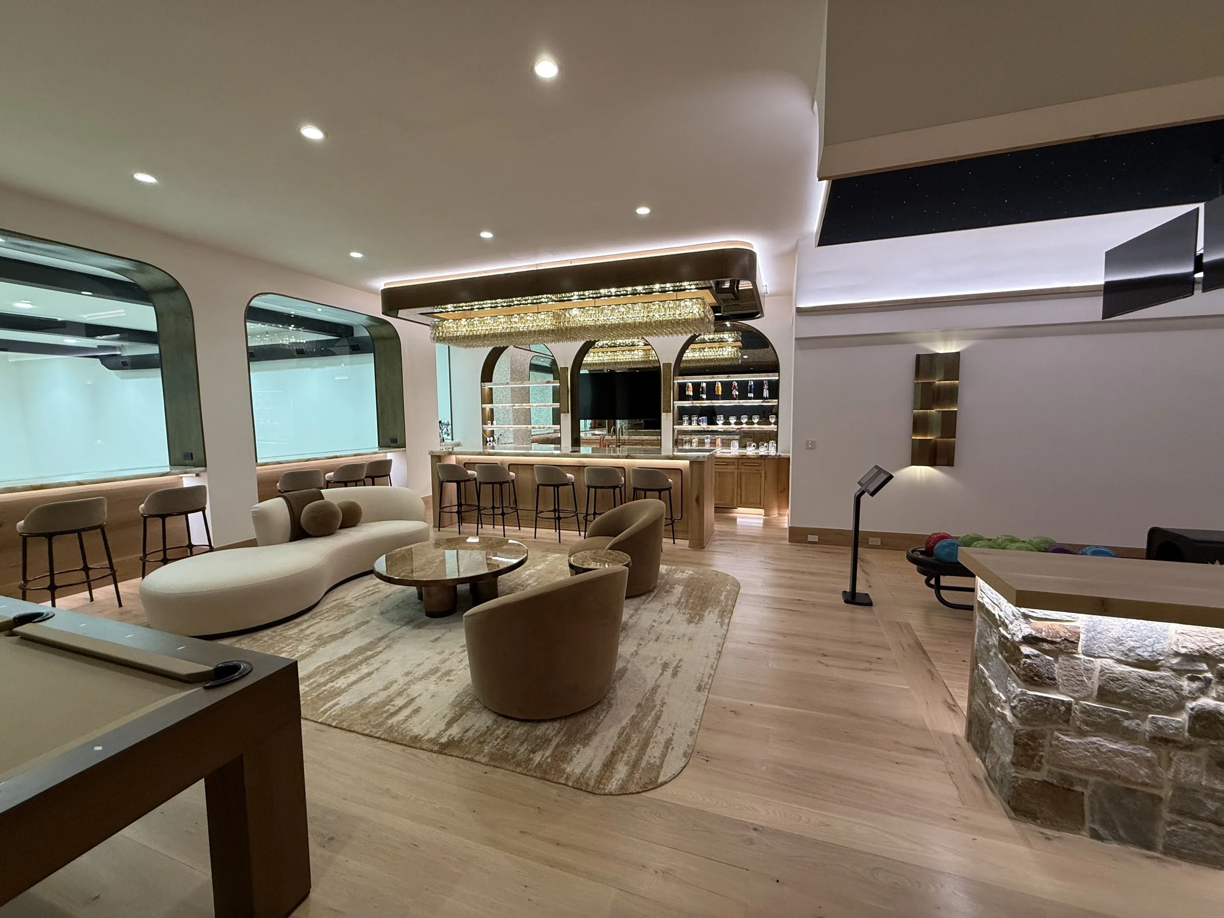 Interior of a modern lounge or entertainment area with seating, a bar, decorative lighting, and a large TV screen.