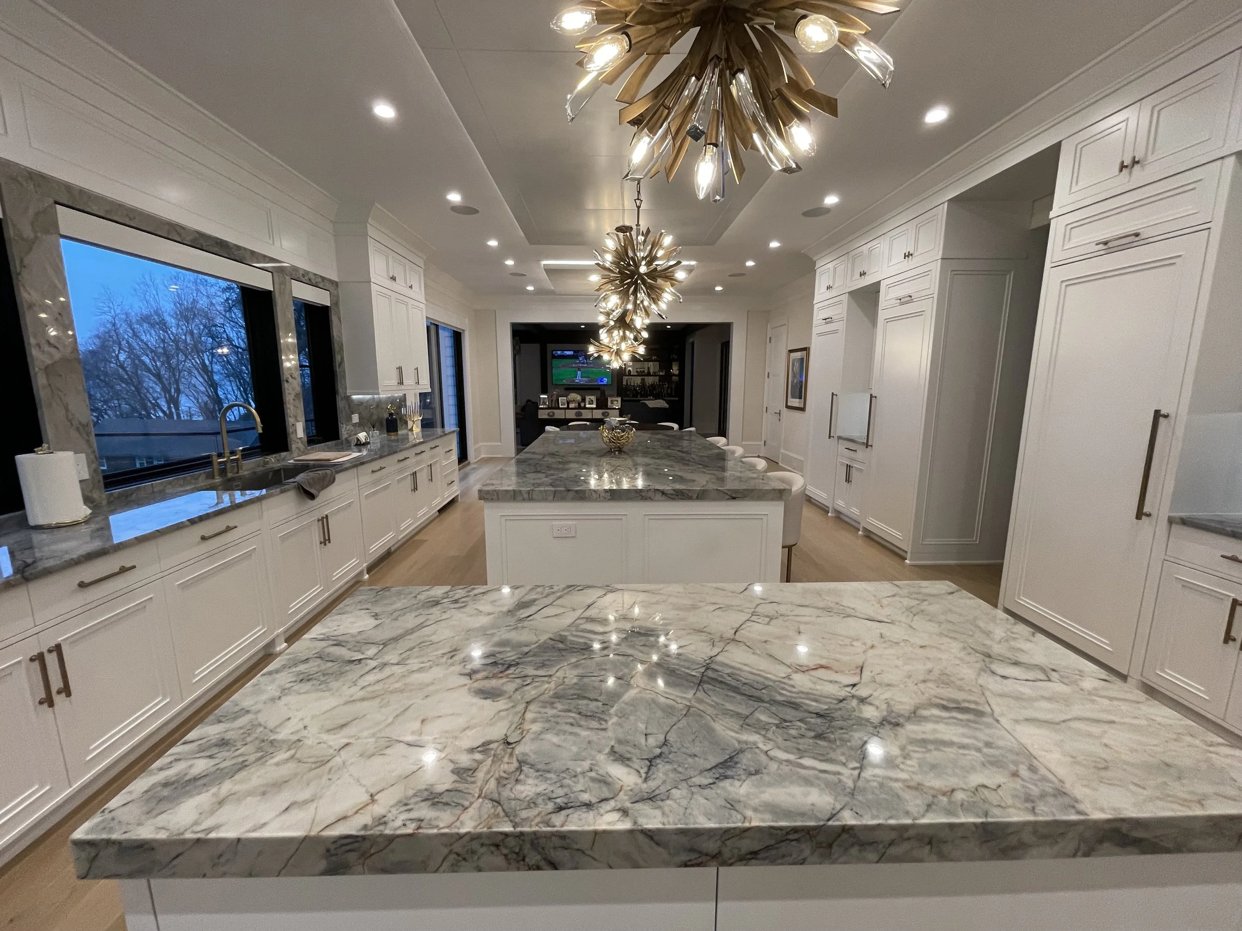 View of a modern kitchen with white cabinets, marble countertops, a large central island, and two elaborate ceiling light fixtures. A window shows a winter scene with bare trees outside.