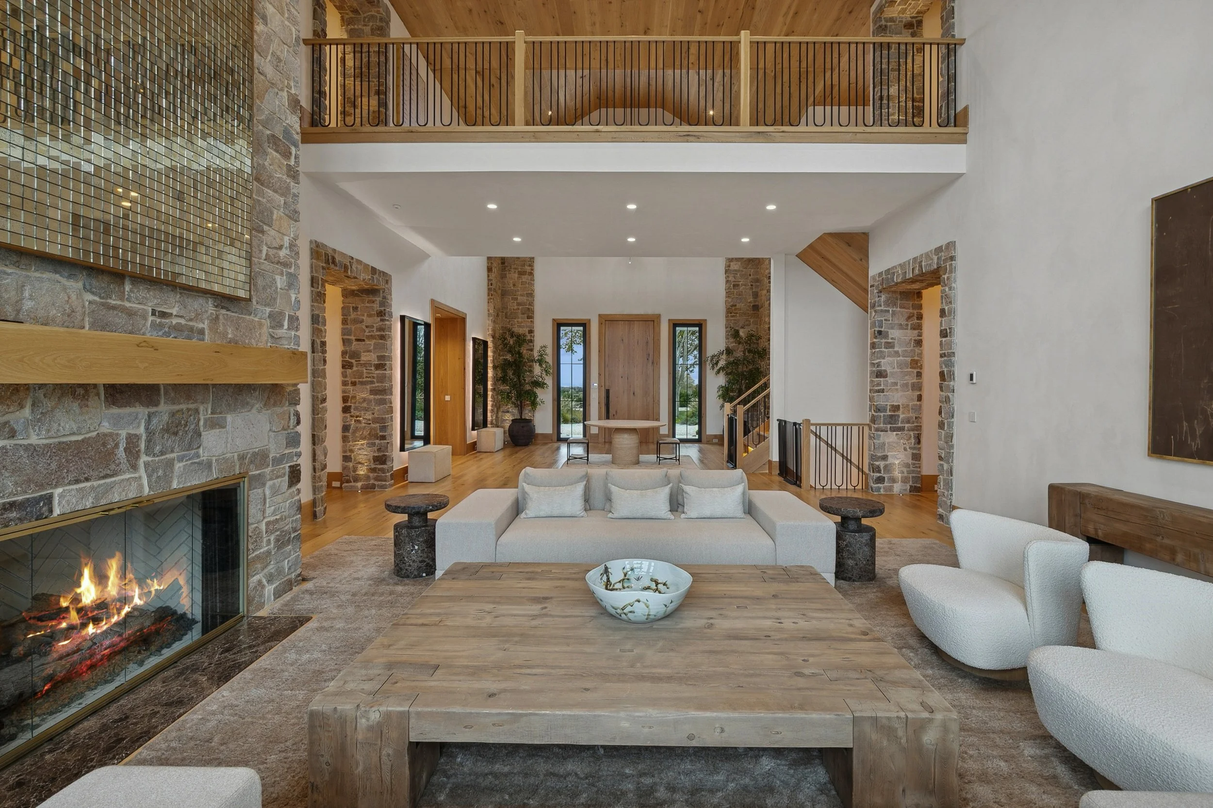 Open-concept living room with stone fireplace, wooden accents, modern furniture, and large windows.