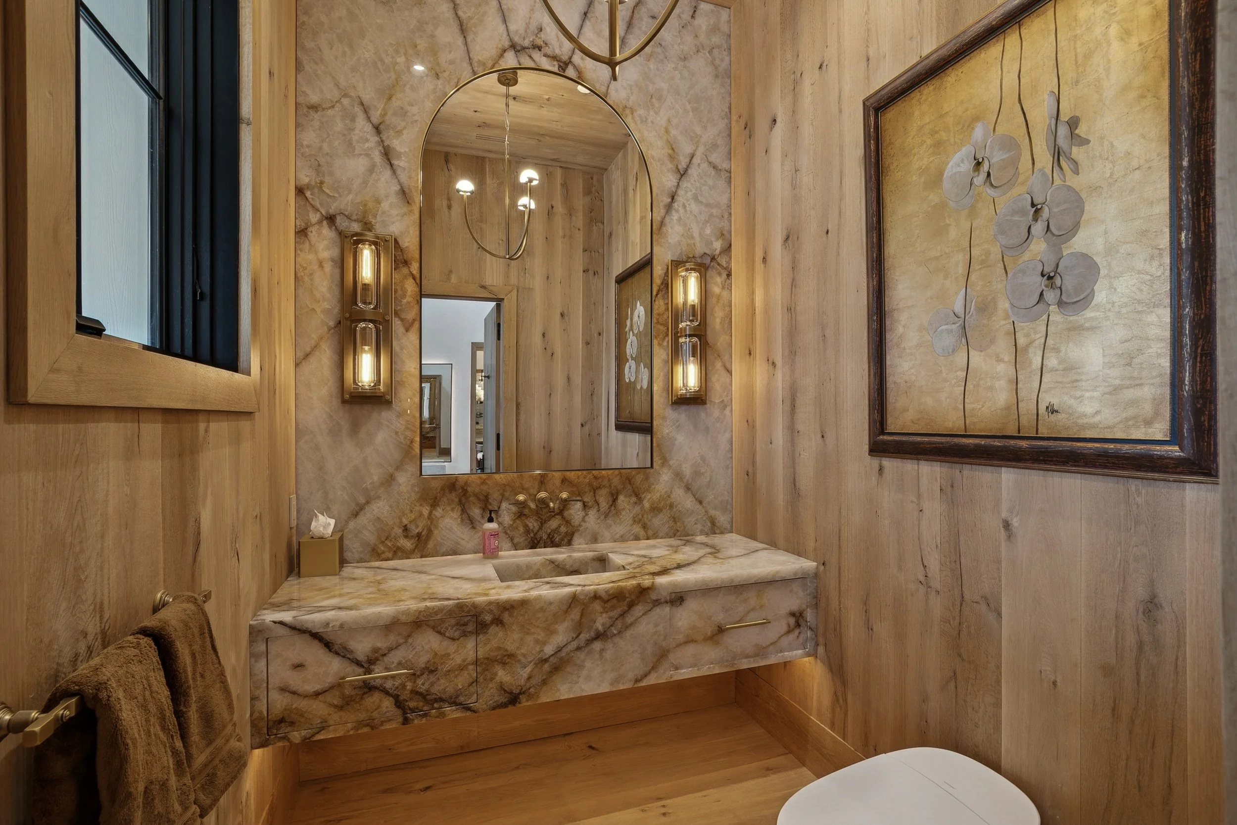 A bathroom with wood-paneled walls and a marble countertop with a sink. There is a large mirror and wall-mounted light fixtures. A framed floral artwork hangs on the wall, and towels are hanging on a towel bar.
