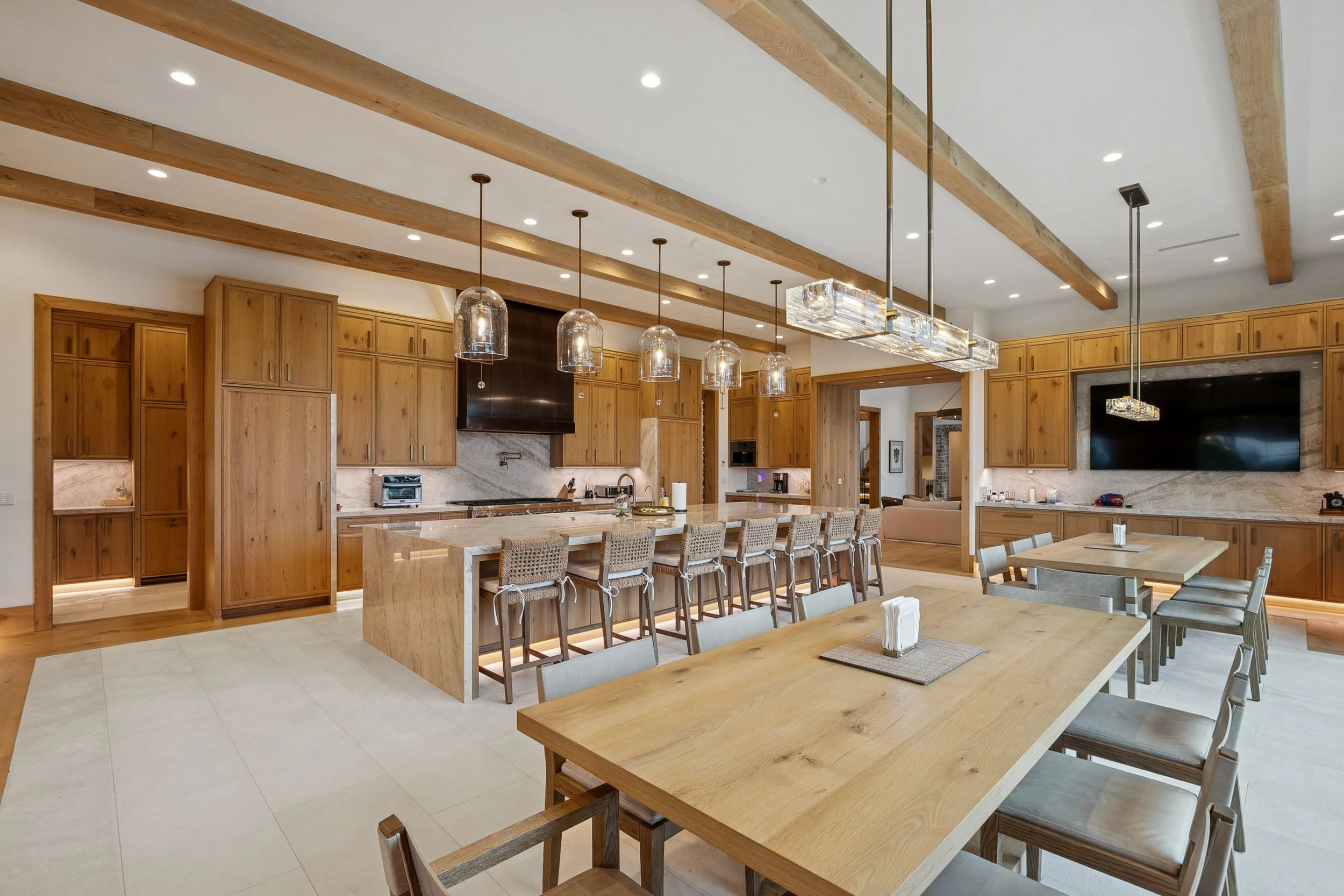 Modern kitchen with wooden cabinets, granite countertops, and a large island with barstools. The ceiling has wooden beams and hanging pendant lights. There is a flat-screen TV on the wall.