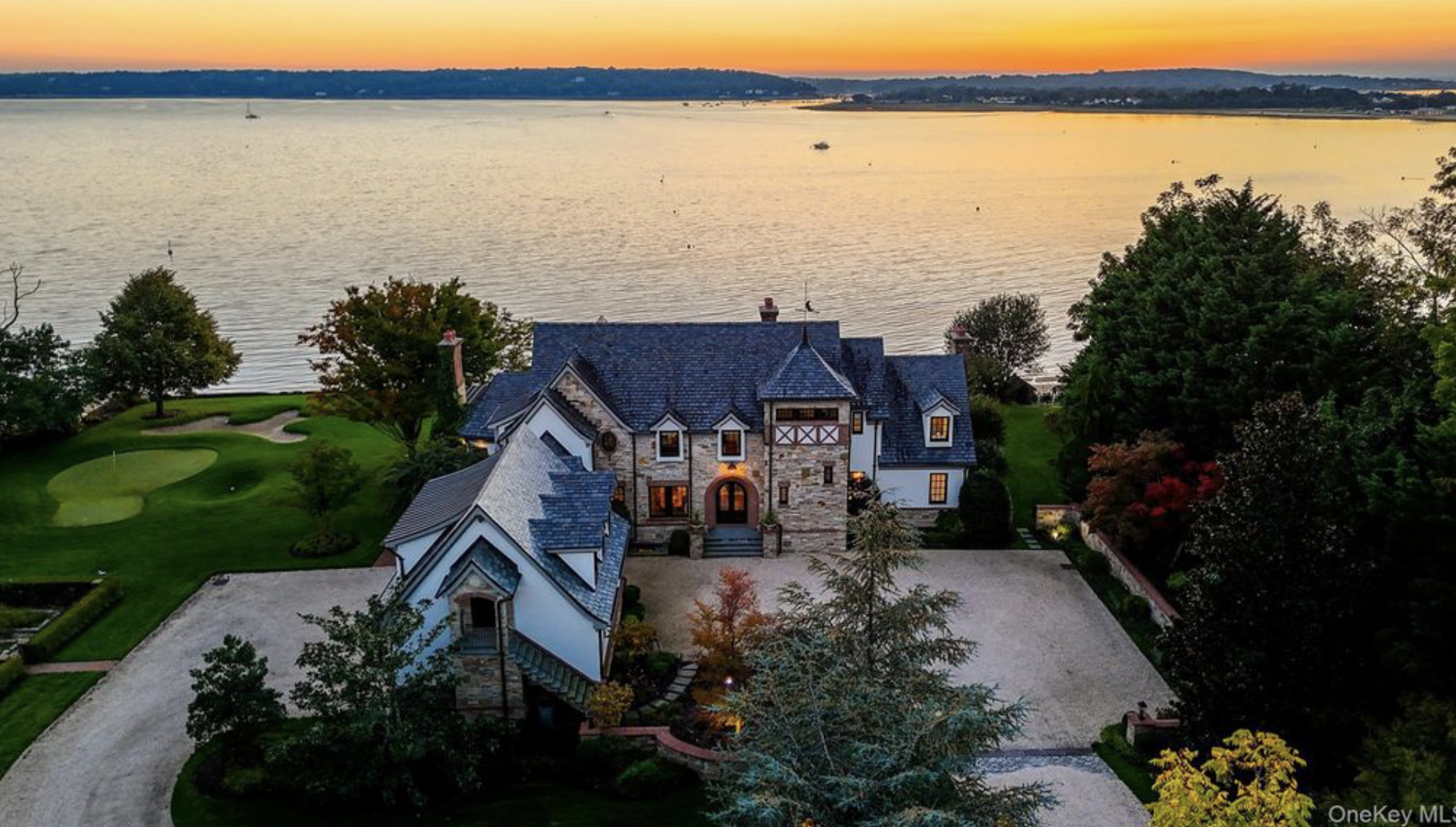 A large, elegant house with stone and white exterior, surrounded by lush trees and a well-maintained lawn. In the background, there is a body of water with a boat, under a vibrant sunset sky with shades of orange and yellow.