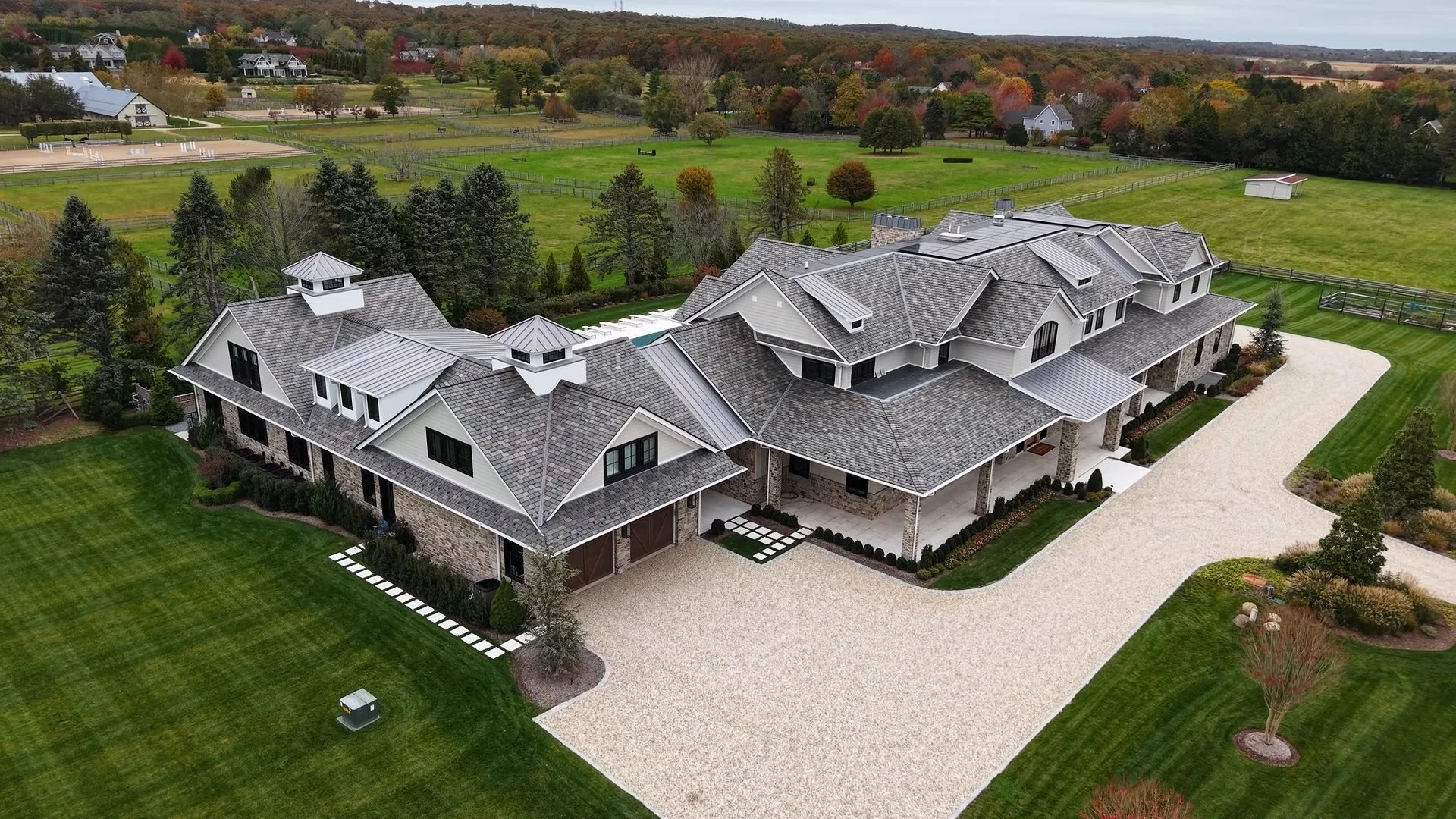 An aerial view of a large, modern house with multiple rooflines, stone and white siding, surrounded by well-maintained lawn and landscape. In the background, expansive fields, trees, and neighboring homes are visible.