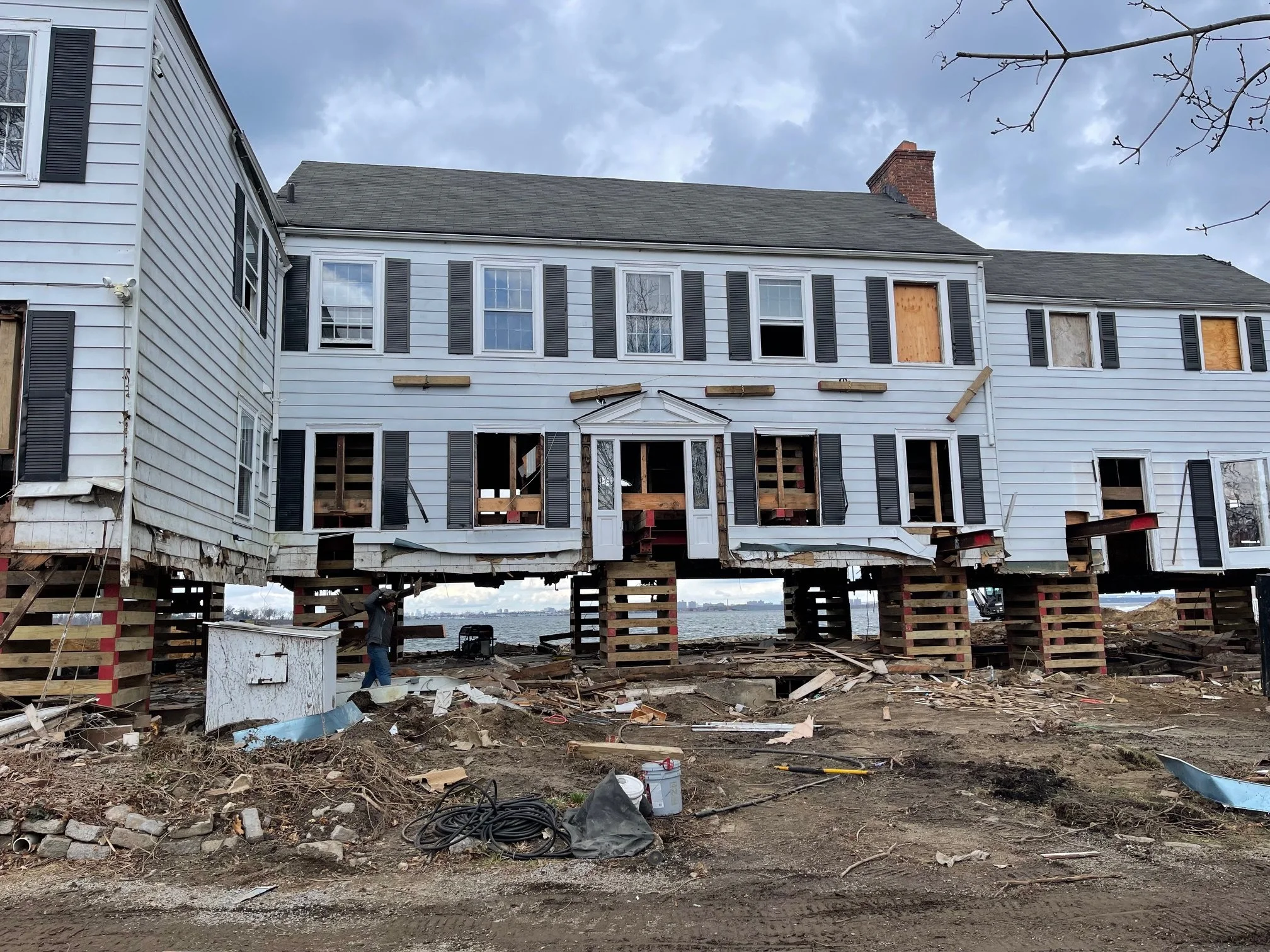 A house under renovation with the lower section demolished, showing the structure's foundation and support beams. The upper part of the house is intact with white siding and black shutters. Construction materials and debris are scattered on the ground, with cloudy skies overhead.