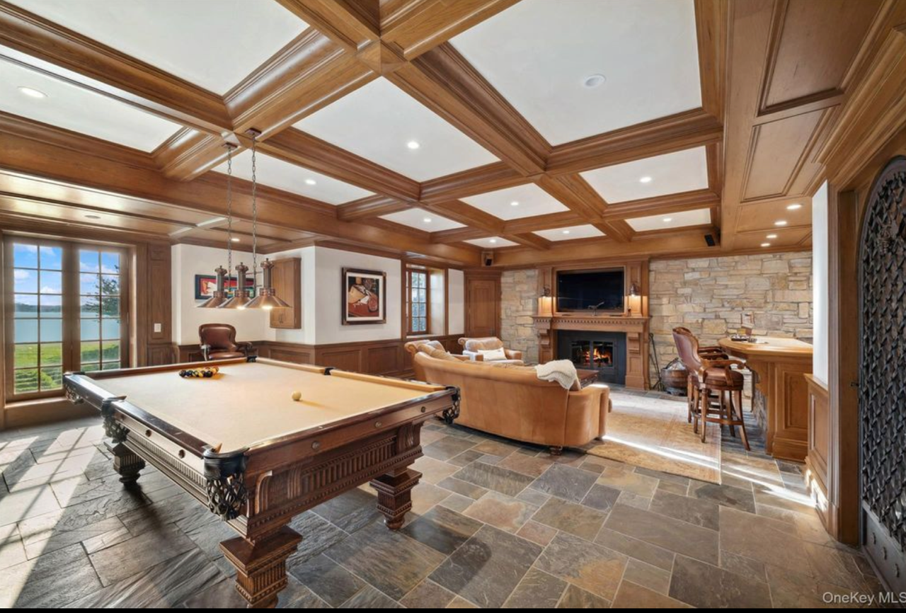 Interior of a cozy game room with a pool table, leather armchairs, a stone fireplace, a flat-screen TV above the fireplace, wooden paneling, and large windows showing a body of water outside.
