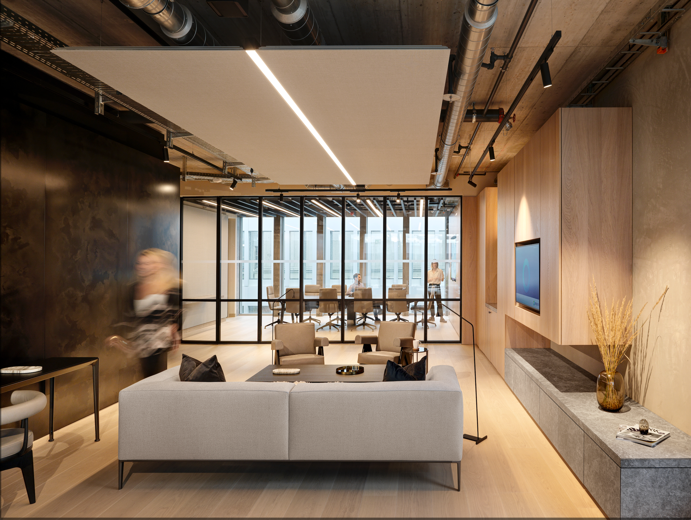 Modern office lounge with a light gray sofa, a black side table with a marble tray, a coffee table with magazines, a wall-mounted TV, and a meeting room with glass walls and six chairs in the background.